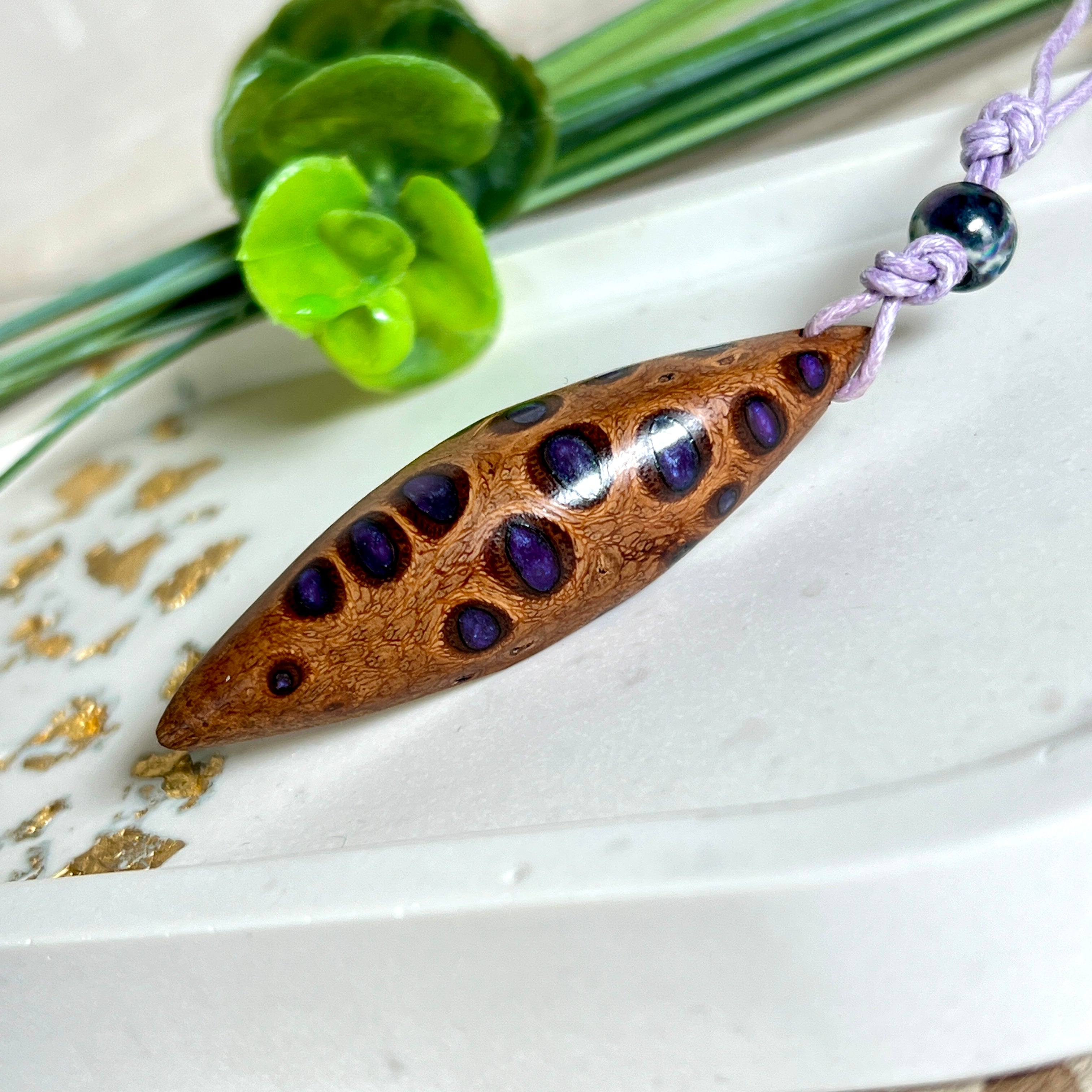 Handgefertigter Anhänger aus einem australischen Banksiazapfen mit eingelassenem metallic-violettem Harz. Der ovale, tropfenförmige Anhänger ist ca. 5,5 cm lang und wird mit einem verstellbaren Baumwollband geliefert. Jedes Stück zeigt die einzigartige natürliche Struktur des Zapfens und schimmert in satten Lilatönen – ein stilvoller Schmuck mit organischem Charakter.