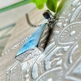 Larimar Anhänger in markanter Dreiecksform, eingefasst in rhodiniertes 925 Sterling Silber, vor strukturiertem Hintergrund mit grünen Blättern – stilvoller Edelsteinschmuck mit karibischem Flair.