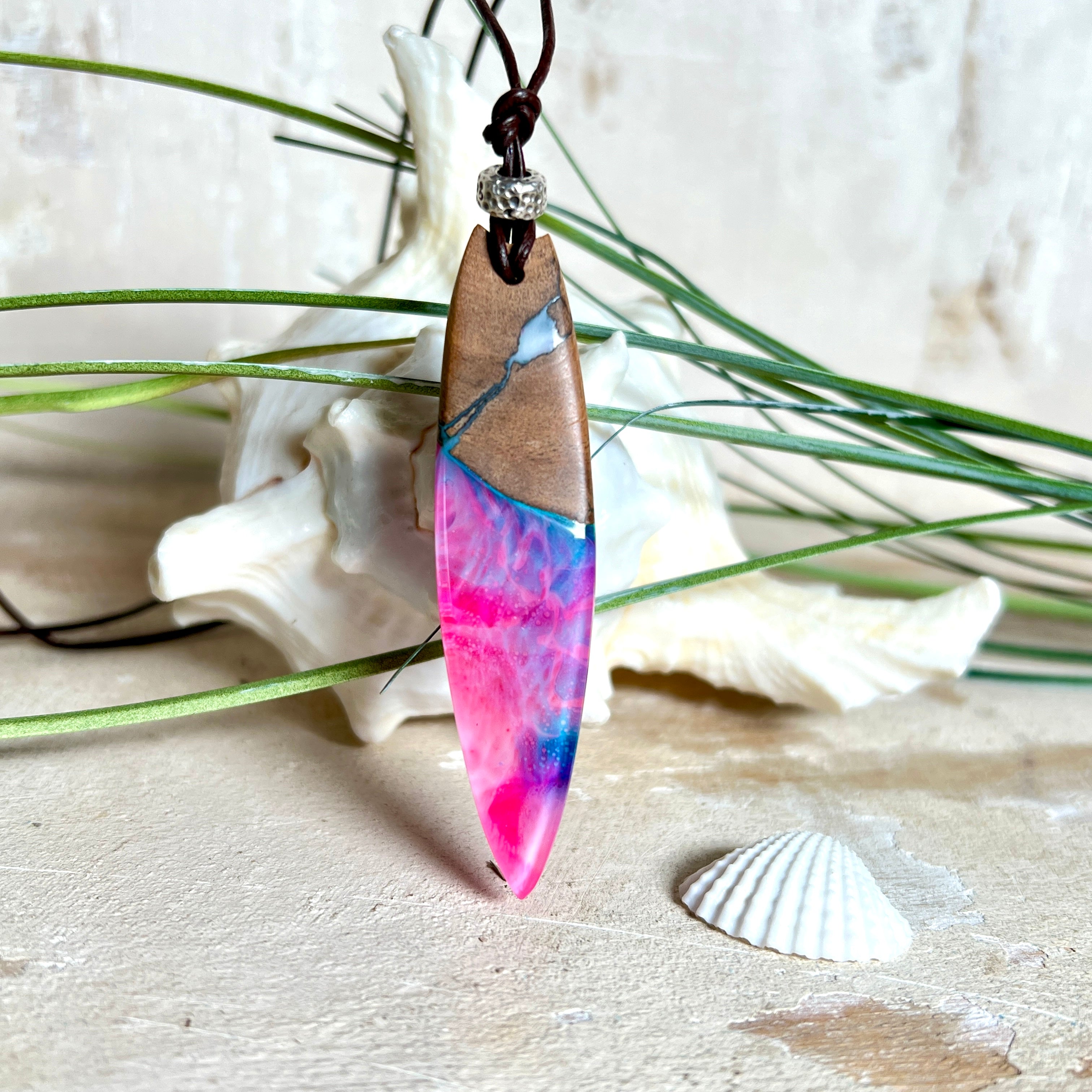 Handgefertigter Surfbrett-Anhänger aus Nussbaumholz und fluoreszierendem Epoxidharz in Pink, Blau und Weiß. Der Anhänger leuchtet zart bei Schwarzlicht und wird an einem verstellbaren Lederband (25–44 cm) mit Holzperlen getragen. Ein maritimes Unikat – ideal für Surfer und Naturliebhaber.