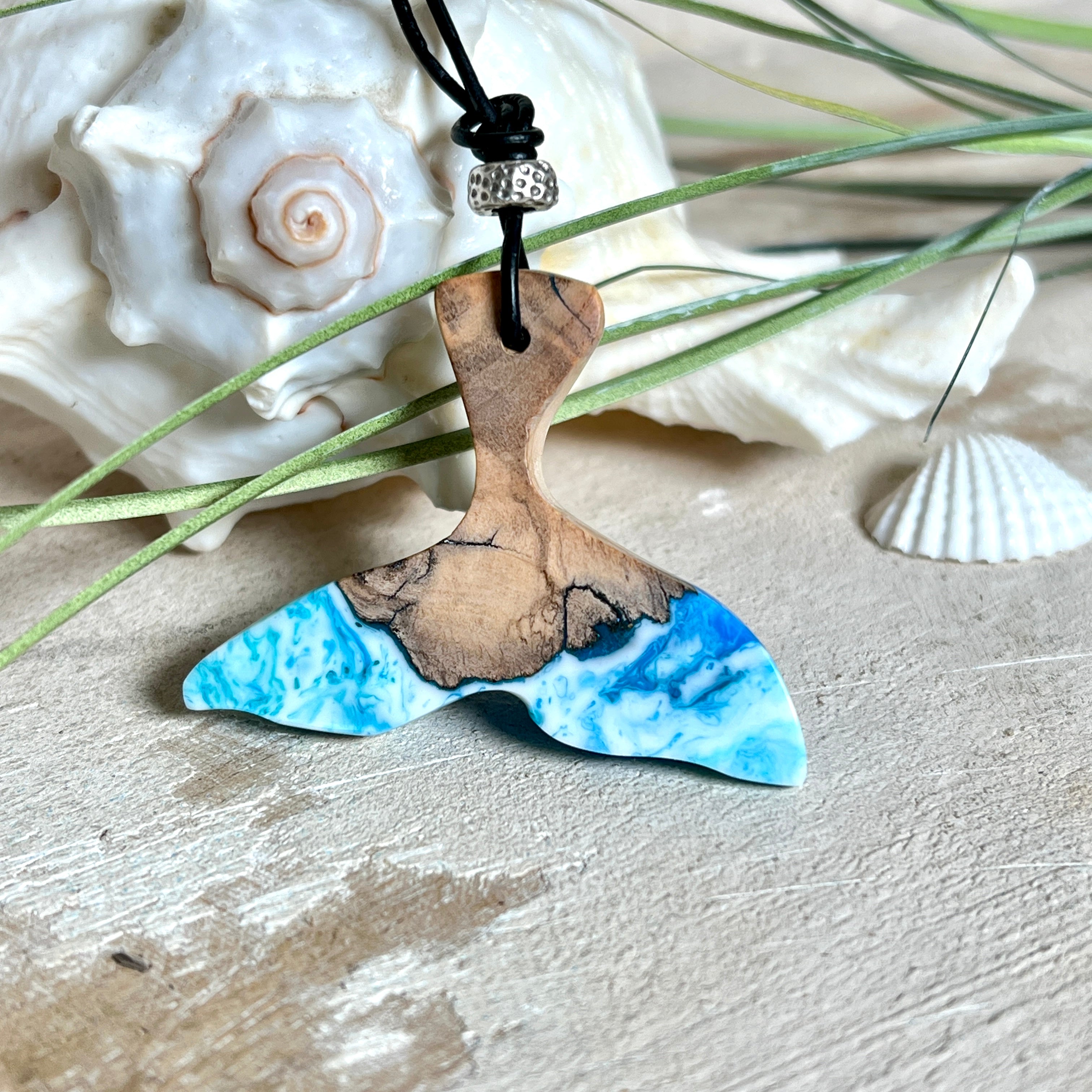 Handgefertigter Walflossen-Anhänger aus Olivenholz und glänzendem Harz in Blau-Grün-Weiß – maritimer Schmuck „Flossenklang“. Der Anhänger erinnert an die Form einer Walflosse und vereint die natürliche Maserung des Holzes mit dem schimmernden Farbspiel des Harzes. Mit verstellbarem schwarzen Lederband und Spacerperle. Ideal für Meeresliebhaber und Naturliebhaber – jedes Stück ein Unikat.