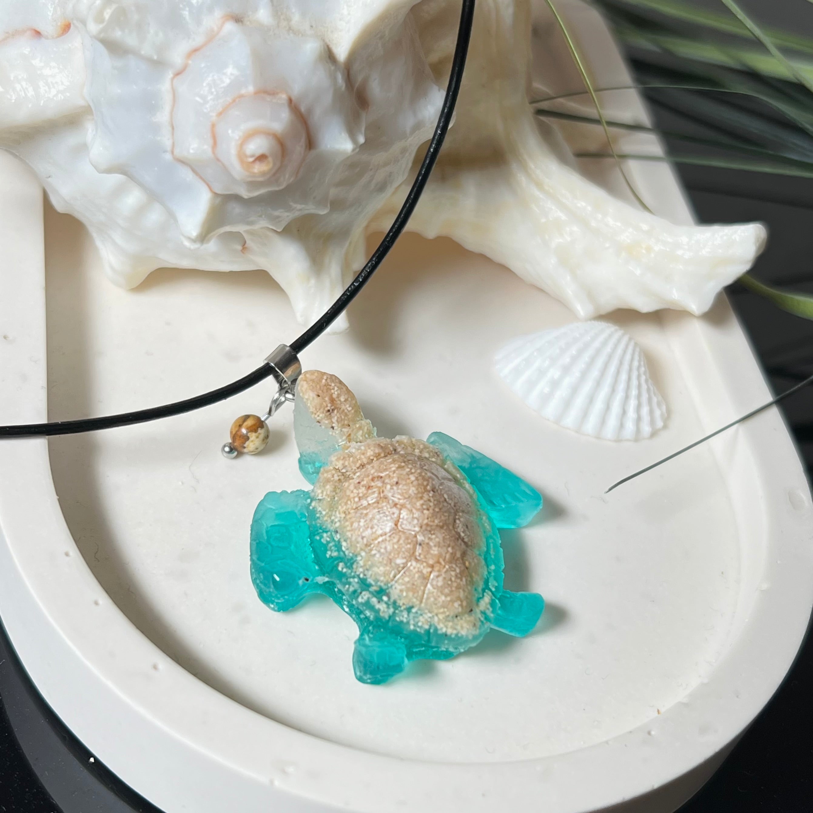 Handgefertigter Schildkröten-Anhänger Turtle Harmony aus echtem Meersand und transparent-türkisfarbenem Harz, kombiniert mit einer kleinen Jaspisperle. Symbol für Weisheit, Schutz und Harmonie. Mit Edelstahl-Anhängerschlaufe und Lederkette (45 + 5 cm). Maritimer Schmuck als Unikat, ideal als liebevolles Geschenk oder stilvolles Accessoire für den Alltag.