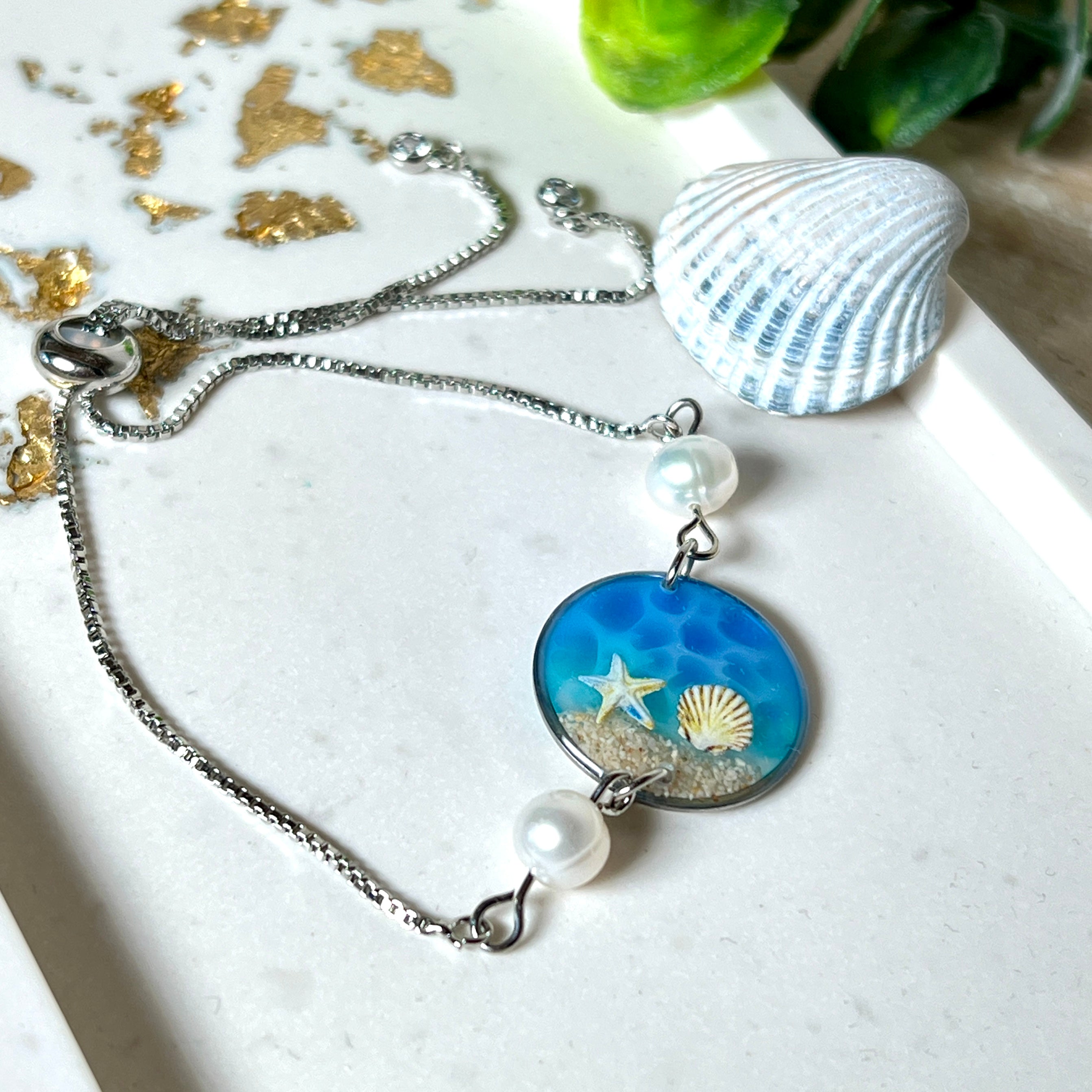 Elegantes Edelstahl-Armband mit rundem Resin-Anhänger in Meeresblau. Im Anhänger liegt echter Meersand als Ufersaum; darüber Sticker-Motive Seestern & Muschel. Zwei feine Süßwasserperlen verzieren das Armband. Verstellbarer Schiebeverschluss . Handgefertigtes Unikat – maritimer Schmuck, der an Wellenrauschen und Strandspaziergänge erinnert.