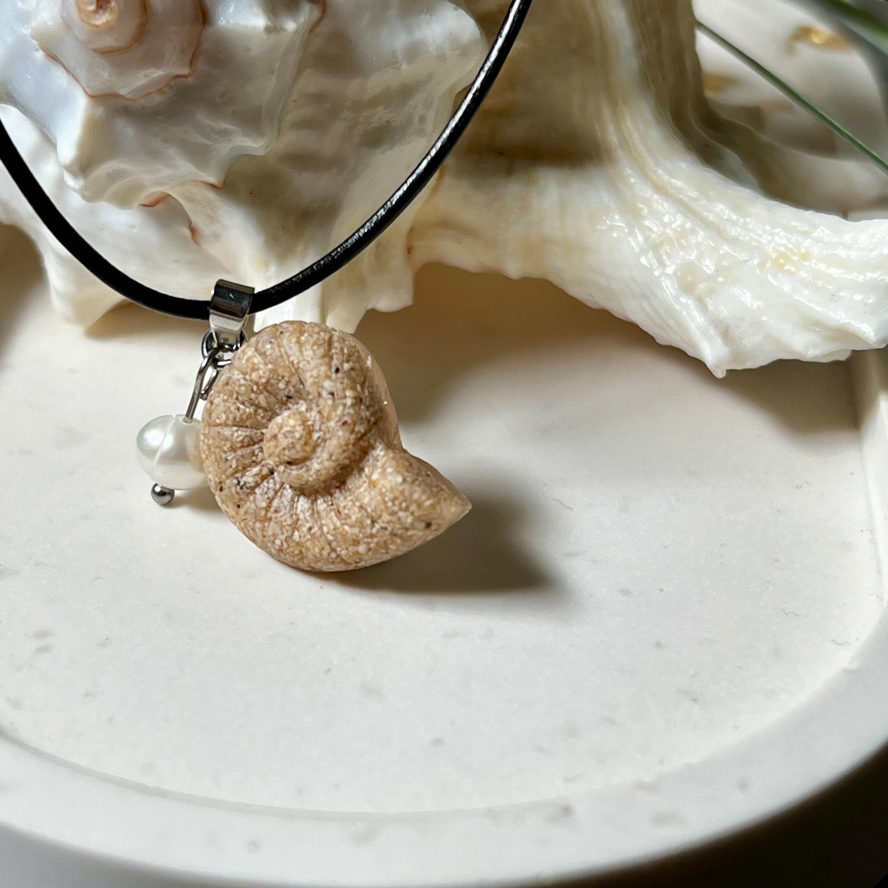 Handgefertigter Ammonit-Anhänger aus echtem Meersand & Harz mit filigraner Edelstahlöse und einer edlen Süßwasserperle. Maritimer Schmuck, inspiriert von der Kraft und Schönheit des Ozeans. Jedes Stück ein Unikat. Präsentiert auf einer Muschel, mit Lederkette.
