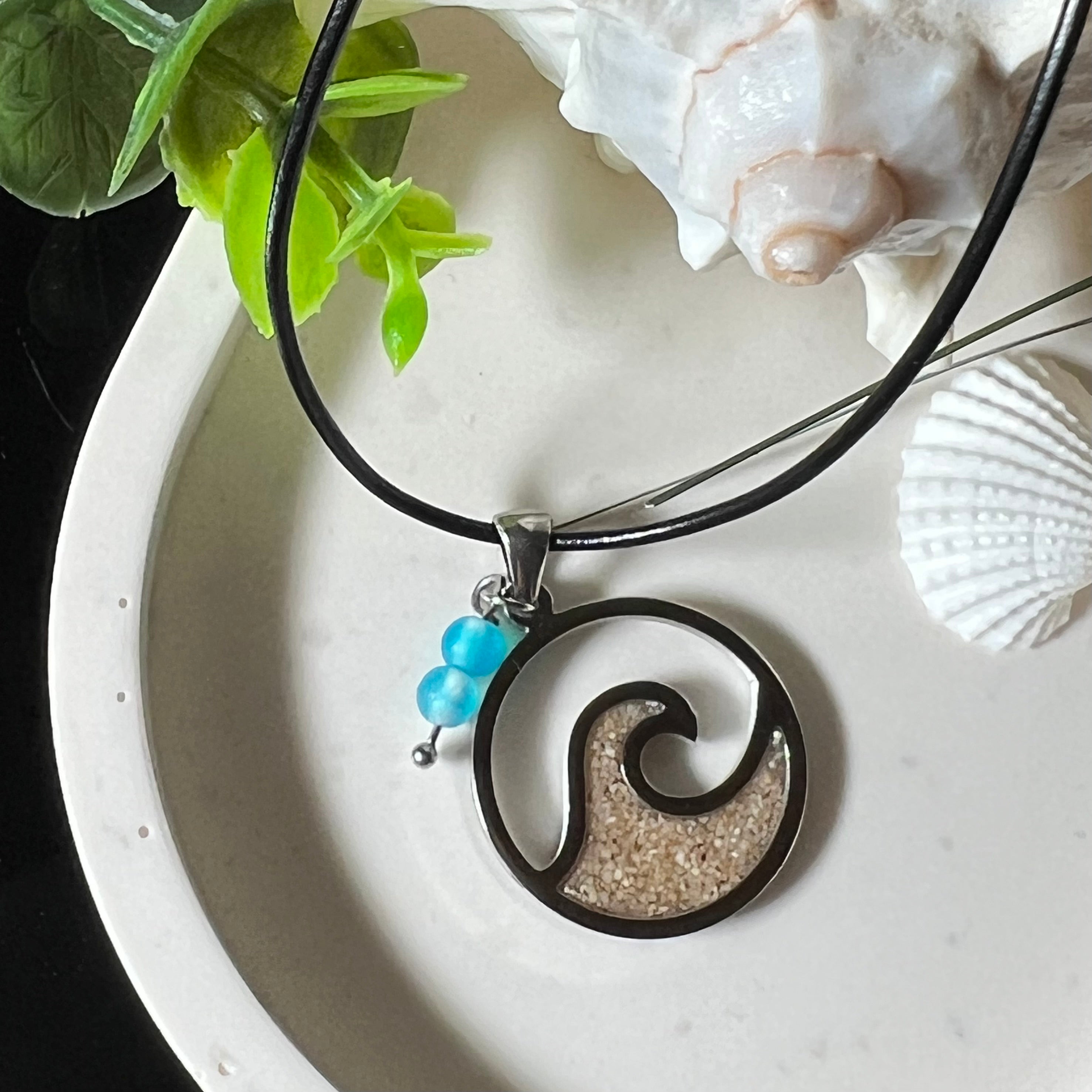 Wave Spirit – runder Edelstahlanhänger mit echter Sandfüllung in Harz eingebettet, inspiriert von einer stilisierten Welle. Verziert mit zwei blauen Achatperlen. Der Anhänger (ca. 2,5 cm Durchmesser) hängt an einer 45 cm langen Lederkette mit Edelstahlverschluss und 5 cm Verlängerungskette. Maritimer Look trifft moderne Eleganz – ein handgefertigtes Schmuckstück für Meerliebhaber