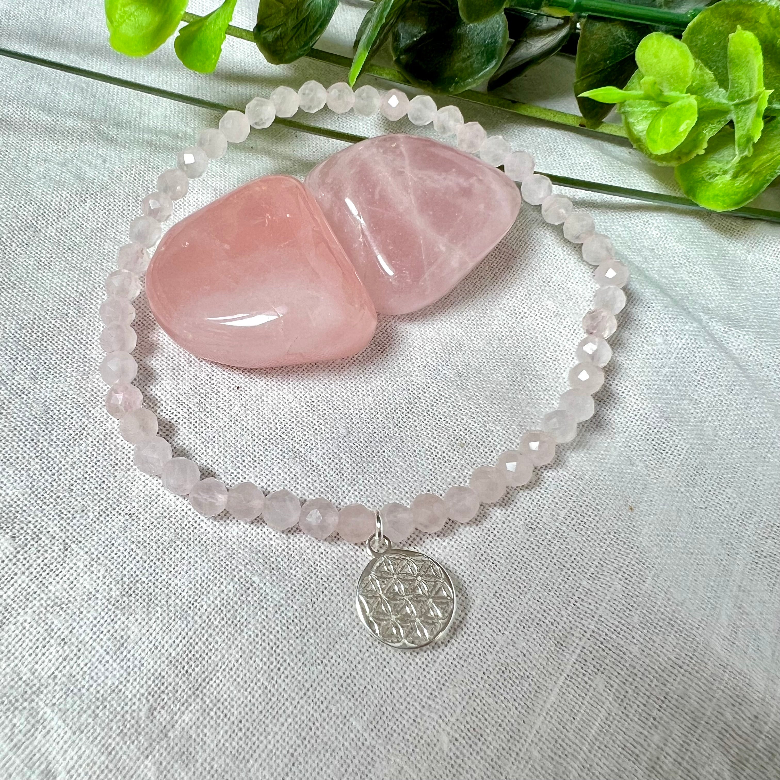 Handgefertigtes Rosenquarz-Armband mit facettierten 4mm Perlen, ergänzt durch einen filigranen Blume-des-Lebens-Anhänger aus 925er Silber. Präsentiert auf hellem Hintergrund mit Rosenquarzsteinen und goldenen Dekoelementen. Spiritueller Schmuck mit harmonisierender Wirkung.
