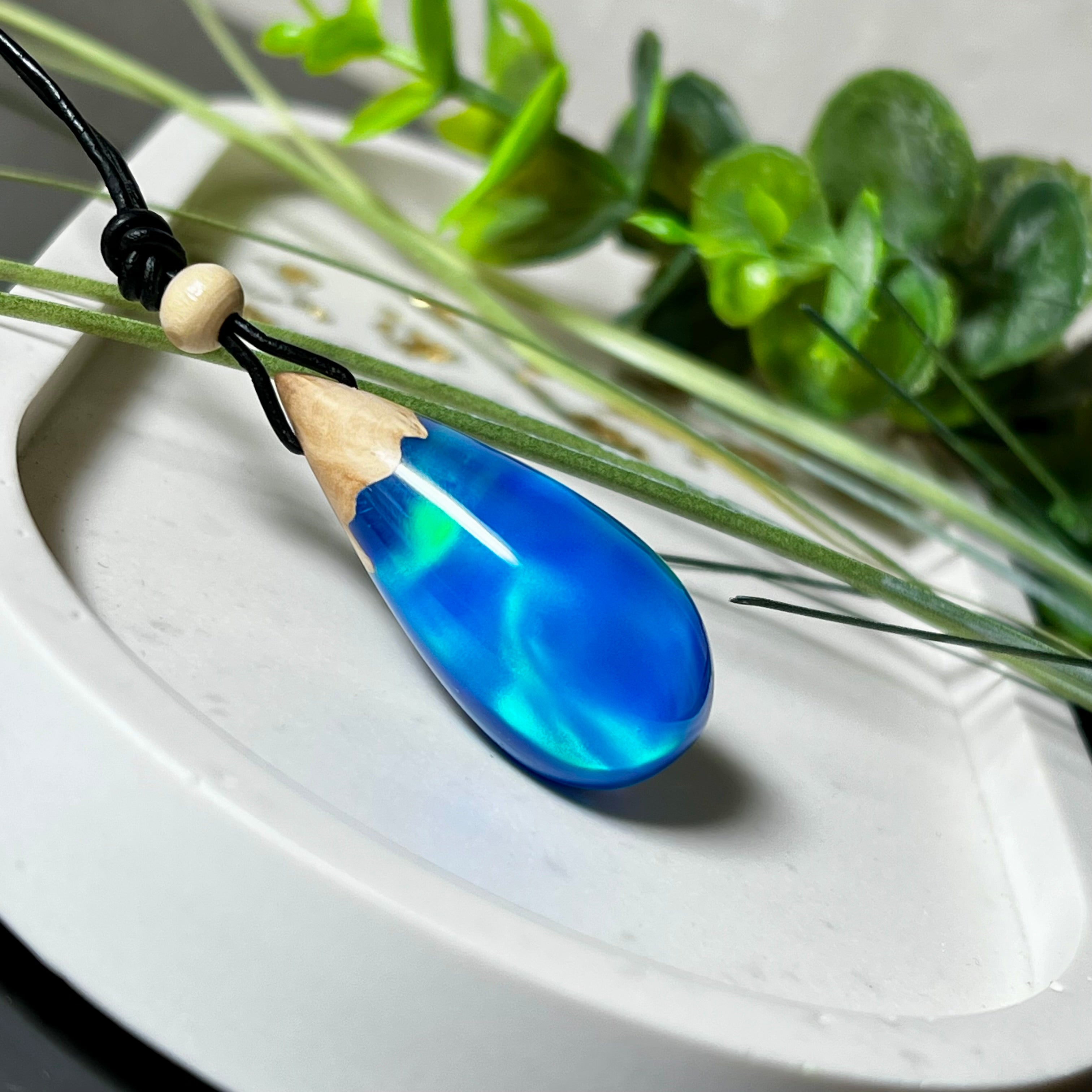 Handgefertigter Tropfenanhänger aus Maserholz und Harz mit synthetischem Opal. Die Farben schimmern je nach Lichteinfall in leuchtendem Blau und Grün – inspiriert von Polarlichtern. Der Anhänger ist ca. 3,9 × 1,8 cm groß und hängt an einem verstellbaren Lederband mit Holzperlen. Jedes Stück ist ein Unikat mit einzigartiger Holzmaserung. Ideal als Geschenk oder stilvoller Begleiter im Alltag.