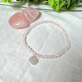 Handgefertigtes Rosenquarz-Armband mit facettierten 4mm Perlen, ergänzt durch einen filigranen Blume-des-Lebens-Anhänger aus 925er Silber. Präsentiert auf hellem Hintergrund mit Rosenquarzsteinen und goldenen Dekoelementen. Spiritueller Schmuck mit harmonisierender Wirkung.