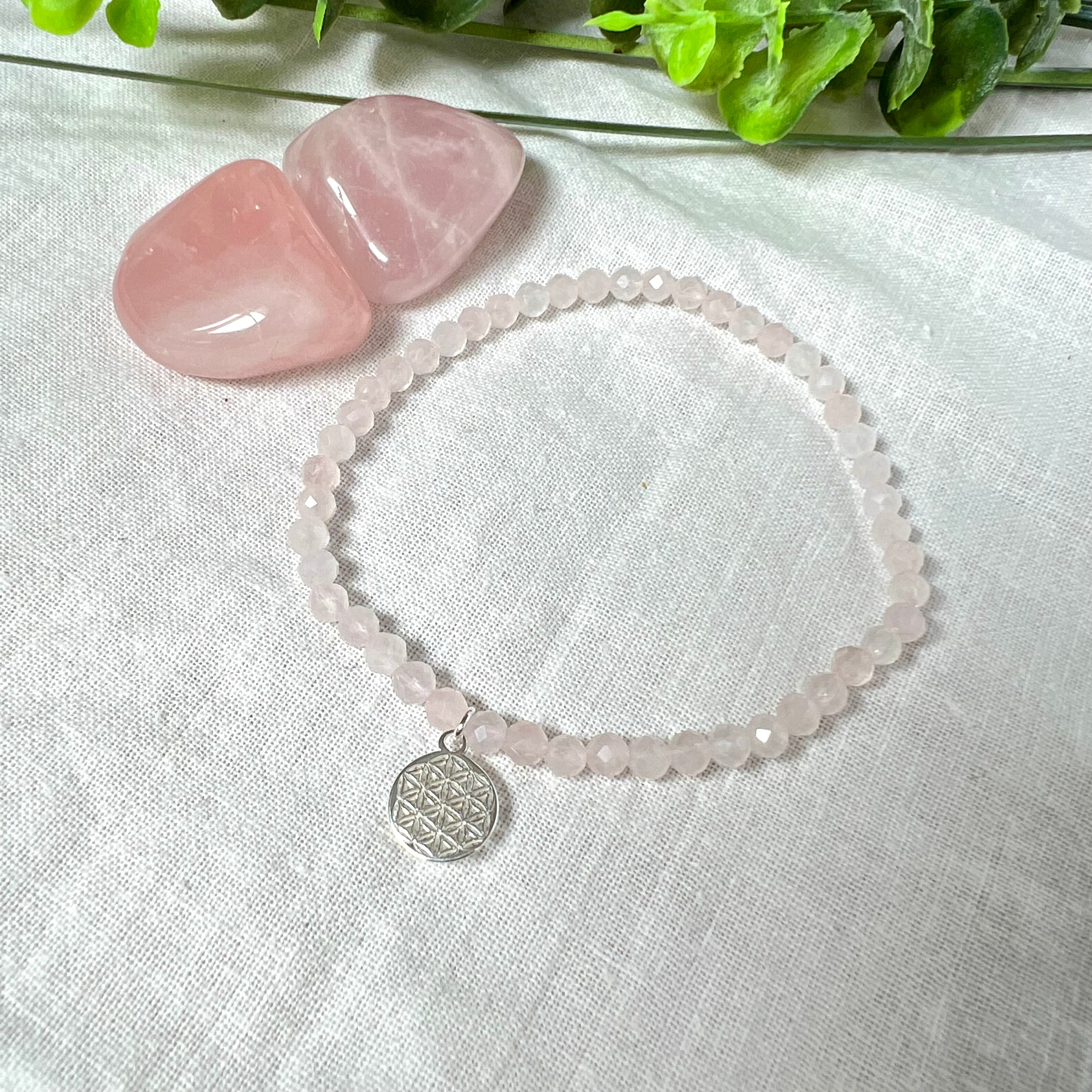 Handgefertigtes Rosenquarz-Armband mit facettierten 4mm Perlen, ergänzt durch einen filigranen Blume-des-Lebens-Anhänger aus 925er Silber. Präsentiert auf hellem Hintergrund mit Rosenquarzsteinen und goldenen Dekoelementen. Spiritueller Schmuck mit harmonisierender Wirkung.
