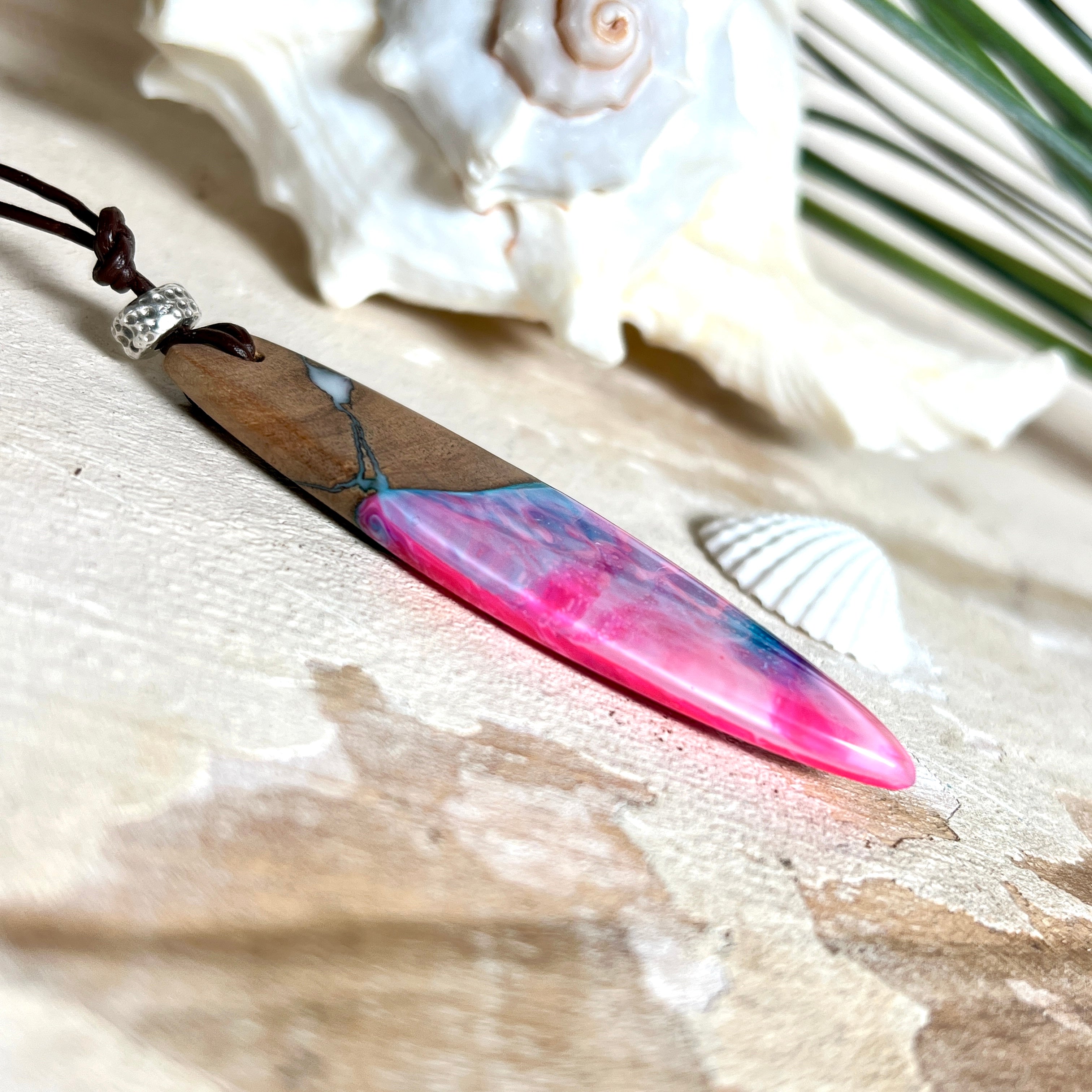 Handgefertigter Surfbrett-Anhänger aus Nussbaumholz und fluoreszierendem Epoxidharz in Pink, Blau und Weiß. Der Anhänger leuchtet zart bei Schwarzlicht und wird an einem verstellbaren Lederband (25–44 cm) mit Holzperlen getragen. Ein maritimes Unikat – ideal für Surfer und Naturliebhaber.
