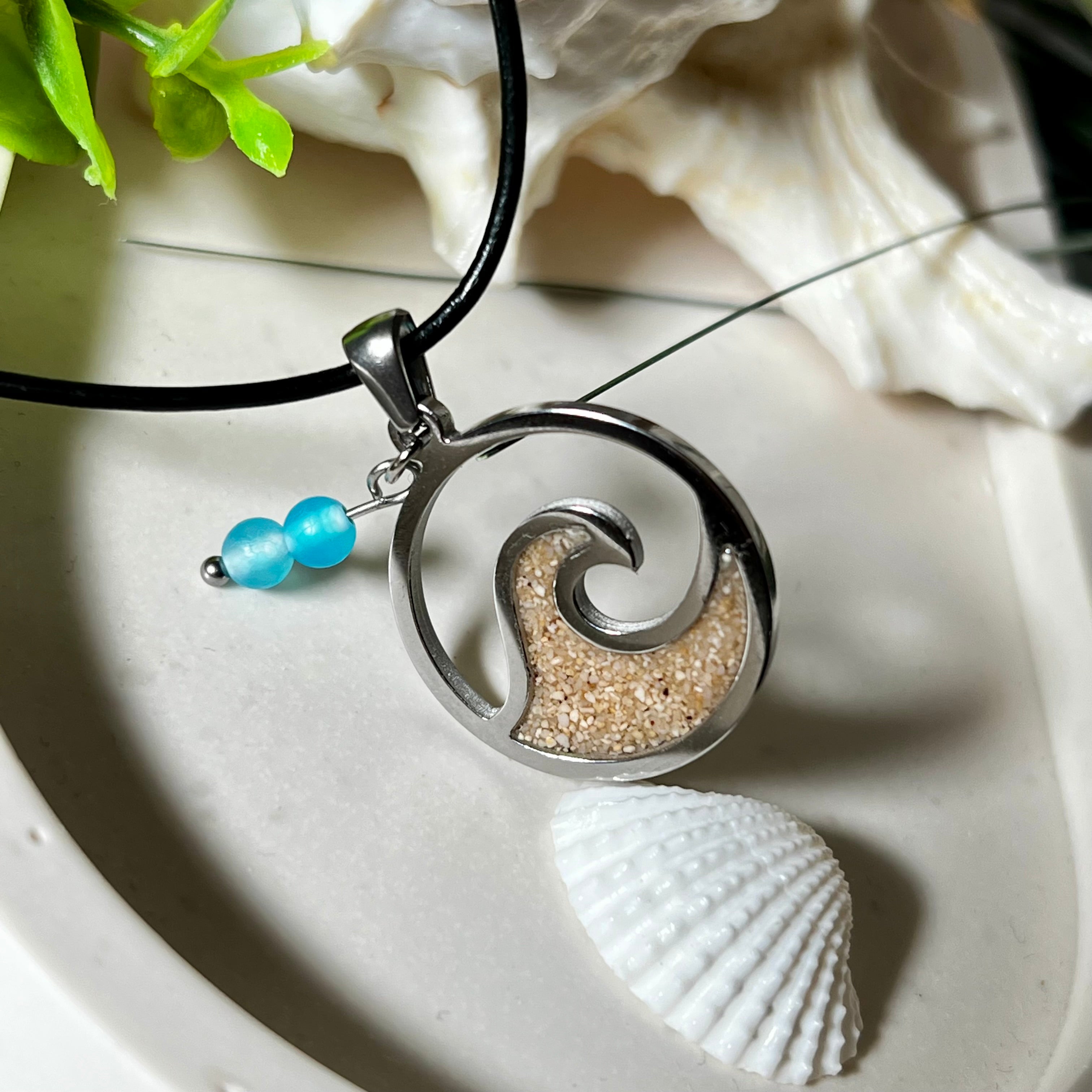 Wave Spirit – runder Edelstahlanhänger mit echter Sandfüllung in Harz eingebettet, inspiriert von einer stilisierten Welle. Verziert mit zwei blauen Achatperlen. Der Anhänger (ca. 2,5 cm Durchmesser) hängt an einer 45 cm langen Lederkette mit Edelstahlverschluss und 5 cm Verlängerungskette. Maritimer Look trifft moderne Eleganz – ein handgefertigtes Schmuckstück für Meerliebhaber