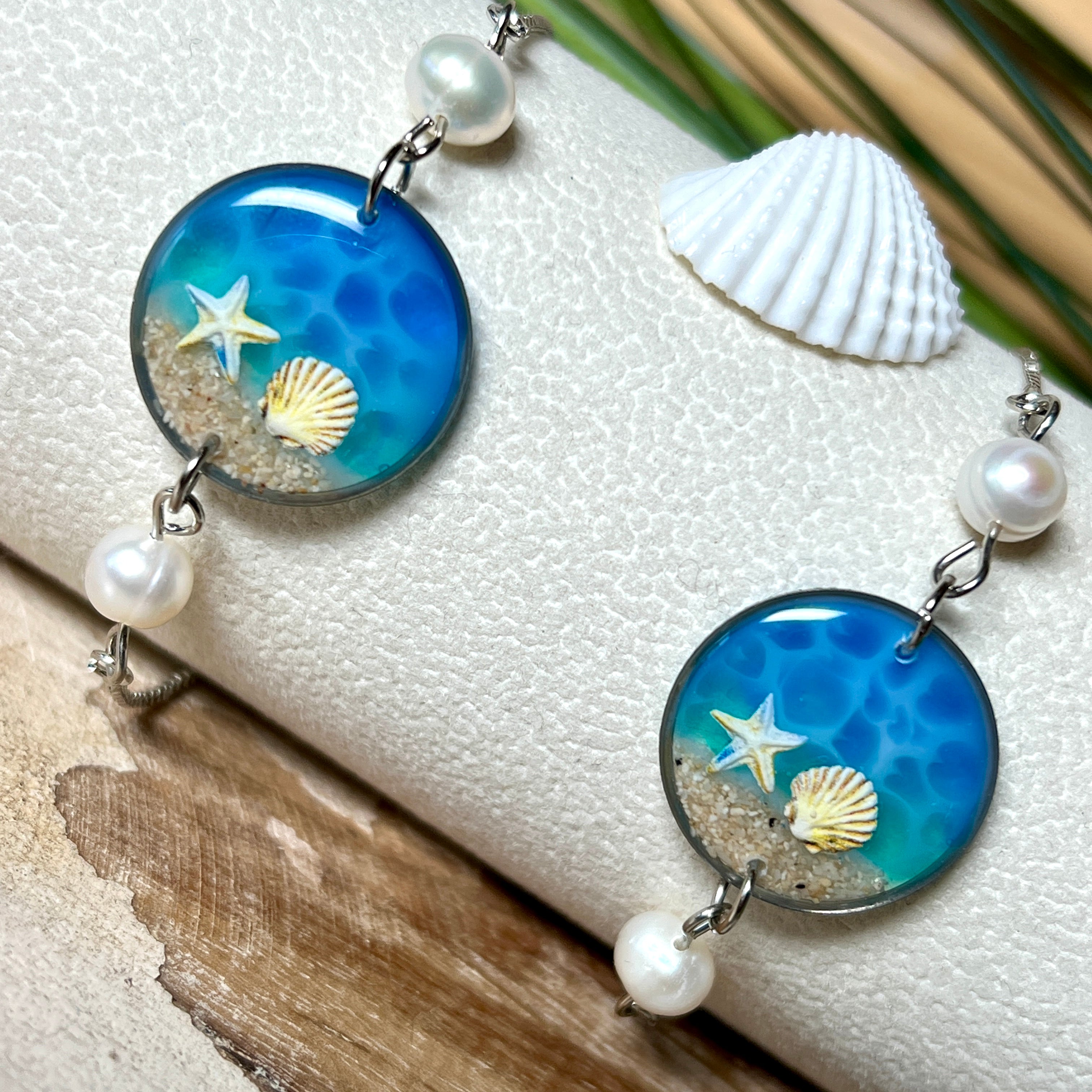Elegantes Edelstahl-Armband mit rundem Resin-Anhänger in Meeresblau. Im Anhänger liegt echter Meersand als Ufersaum; darüber Sticker-Motive Seestern & Muschel. Zwei feine Süßwasserperlen verzieren das Armband. Verstellbarer Schiebeverschluss . Handgefertigtes Unikat – maritimer Schmuck, der an Wellenrauschen und Strandspaziergänge erinnert.