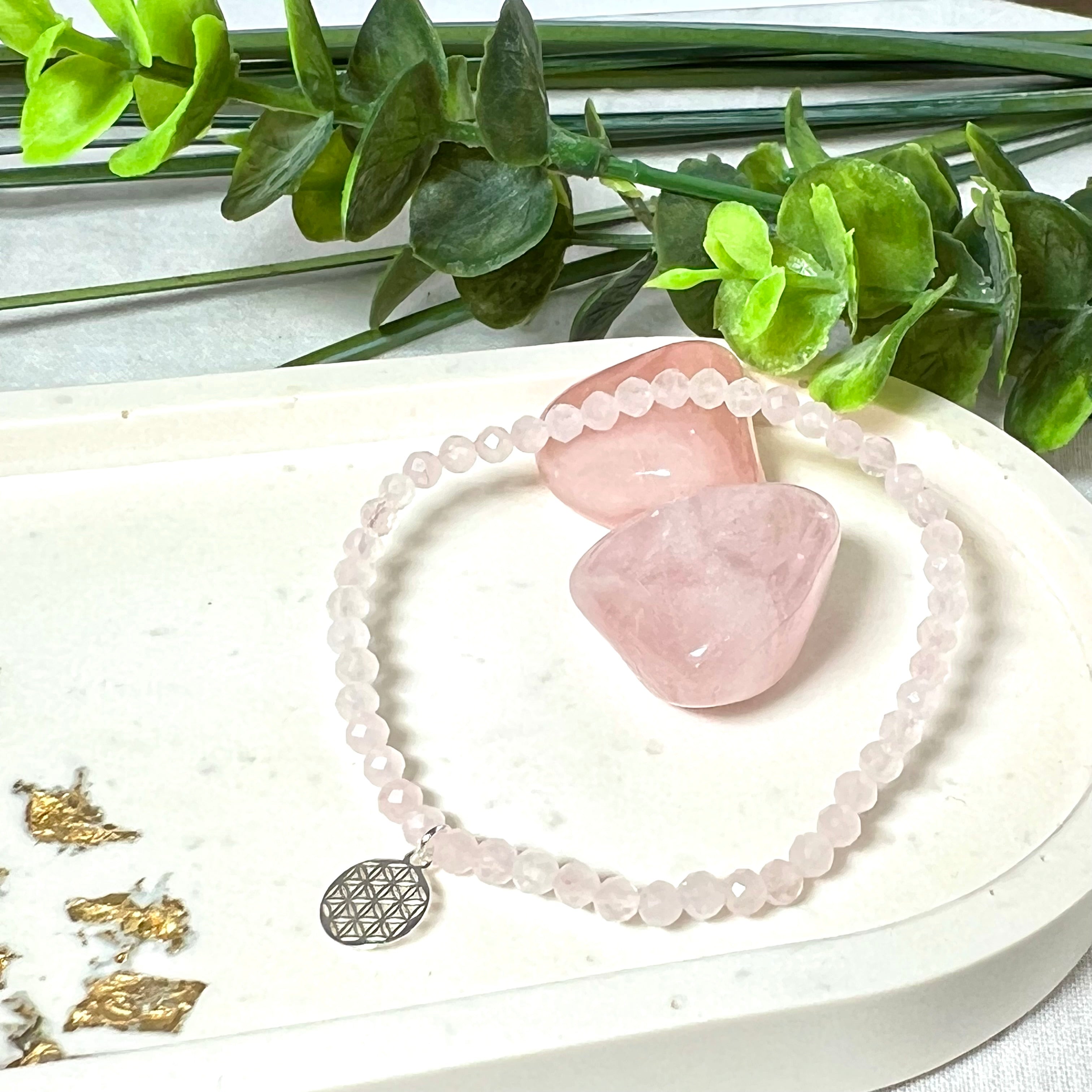 Handgefertigtes Rosenquarz-Armband mit facettierten 4mm Perlen, ergänzt durch einen filigranen Blume-des-Lebens-Anhänger aus 925er Silber. Präsentiert auf hellem Hintergrund mit Rosenquarzsteinen und goldenen Dekoelementen. Spiritueller Schmuck mit harmonisierender Wirkung.