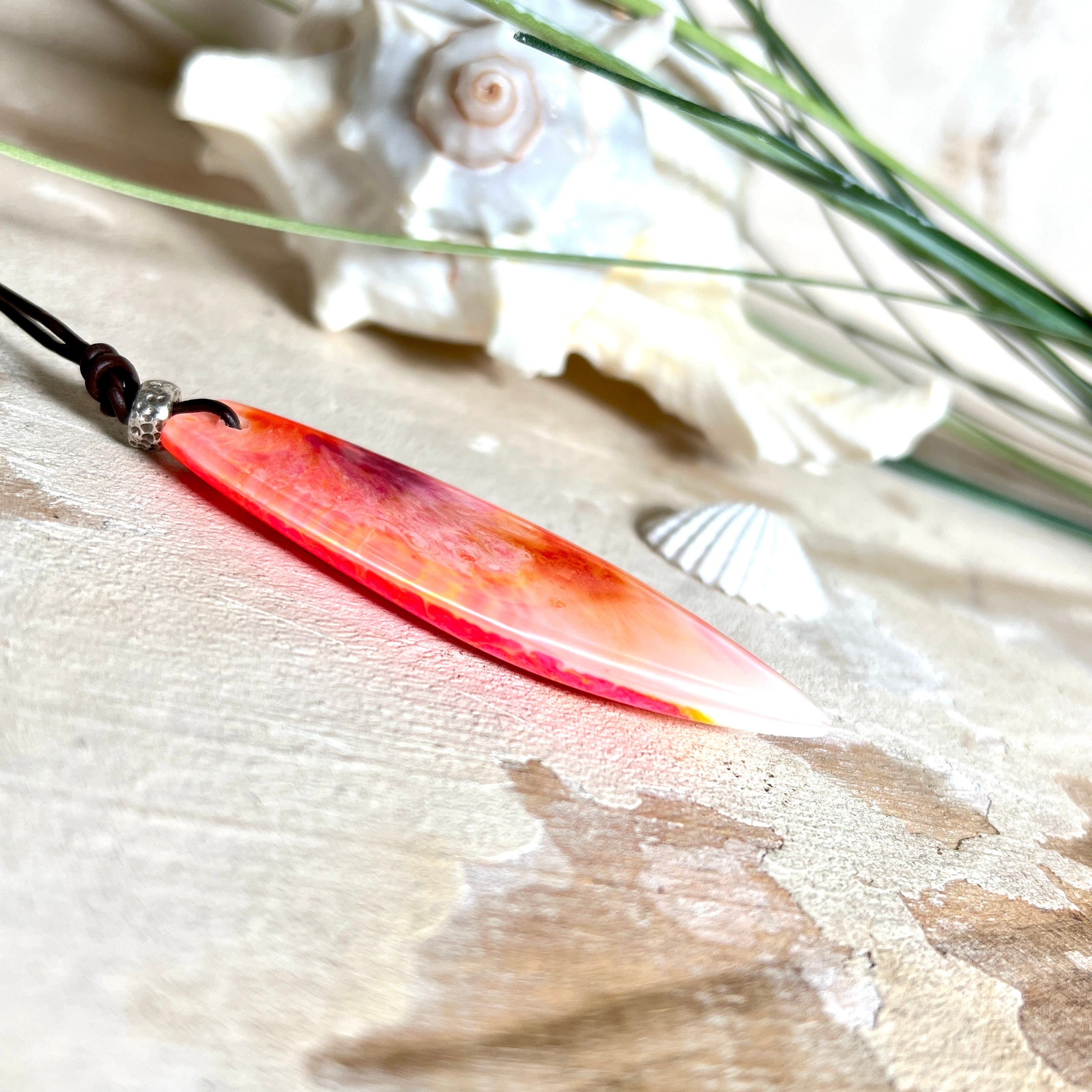 Handgefertigter Surfbrett-Anhänger aus fluoreszierendem Epoxidharz in leuchtenden Orange-, Rot- und Gelbtönen. Leuchtet bei Schwarzlicht. Kombiniert mit verstellbarem Lederband (26–45 cm), verziert mit Spacer- und Holzperlen. Das Design ist inspiriert von der Farbintensität eines Sonnenuntergangs über dem Meer.