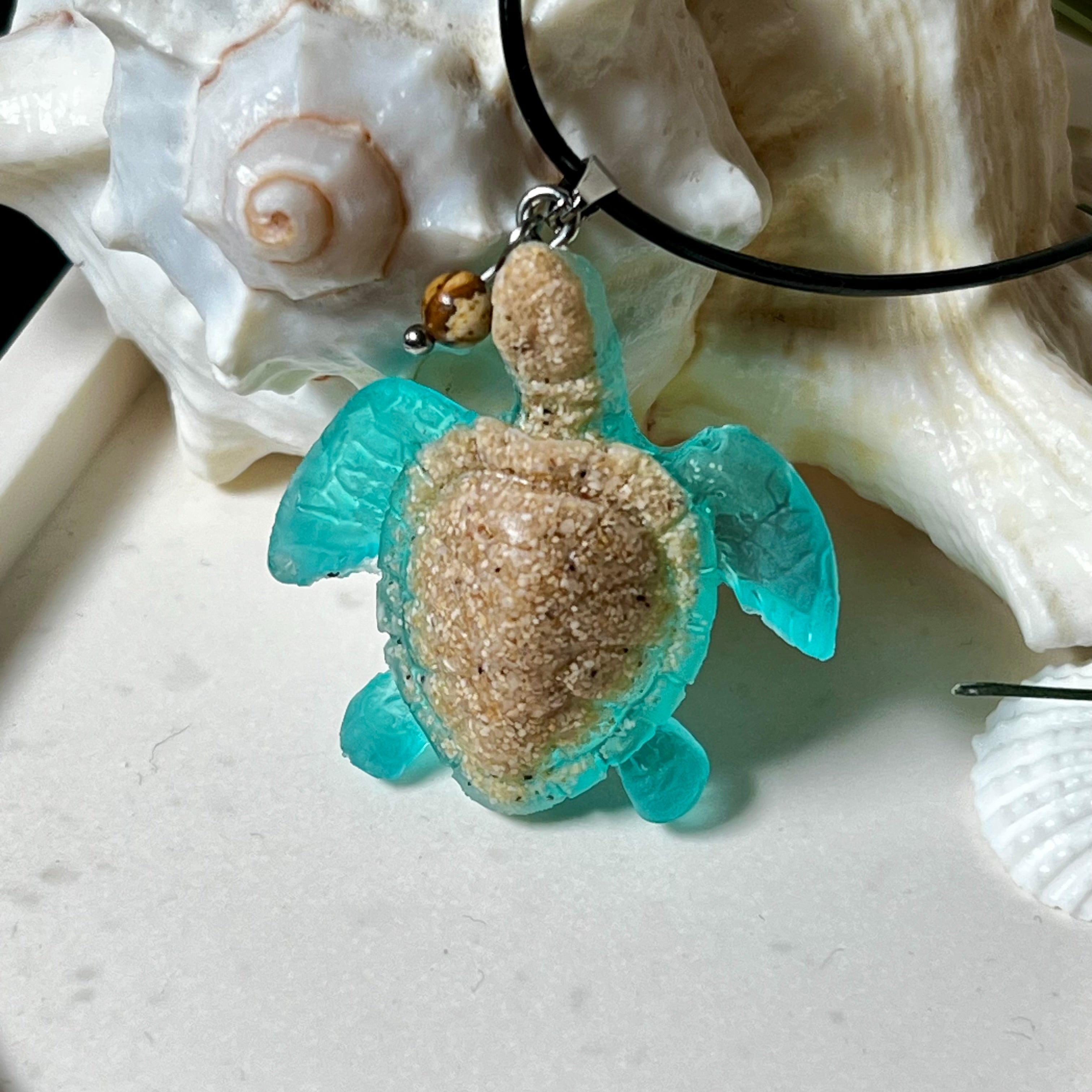 Handgefertigter Schildkröten-Anhänger Turtle Harmony aus echtem Meersand und transparent-türkisfarbenem Harz, kombiniert mit einer kleinen Jaspisperle. Symbol für Weisheit, Schutz und Harmonie. Mit Edelstahl-Anhängerschlaufe und Lederkette (45 + 5 cm). Maritimer Schmuck als Unikat, ideal als liebevolles Geschenk oder stilvolles Accessoire für den Alltag.