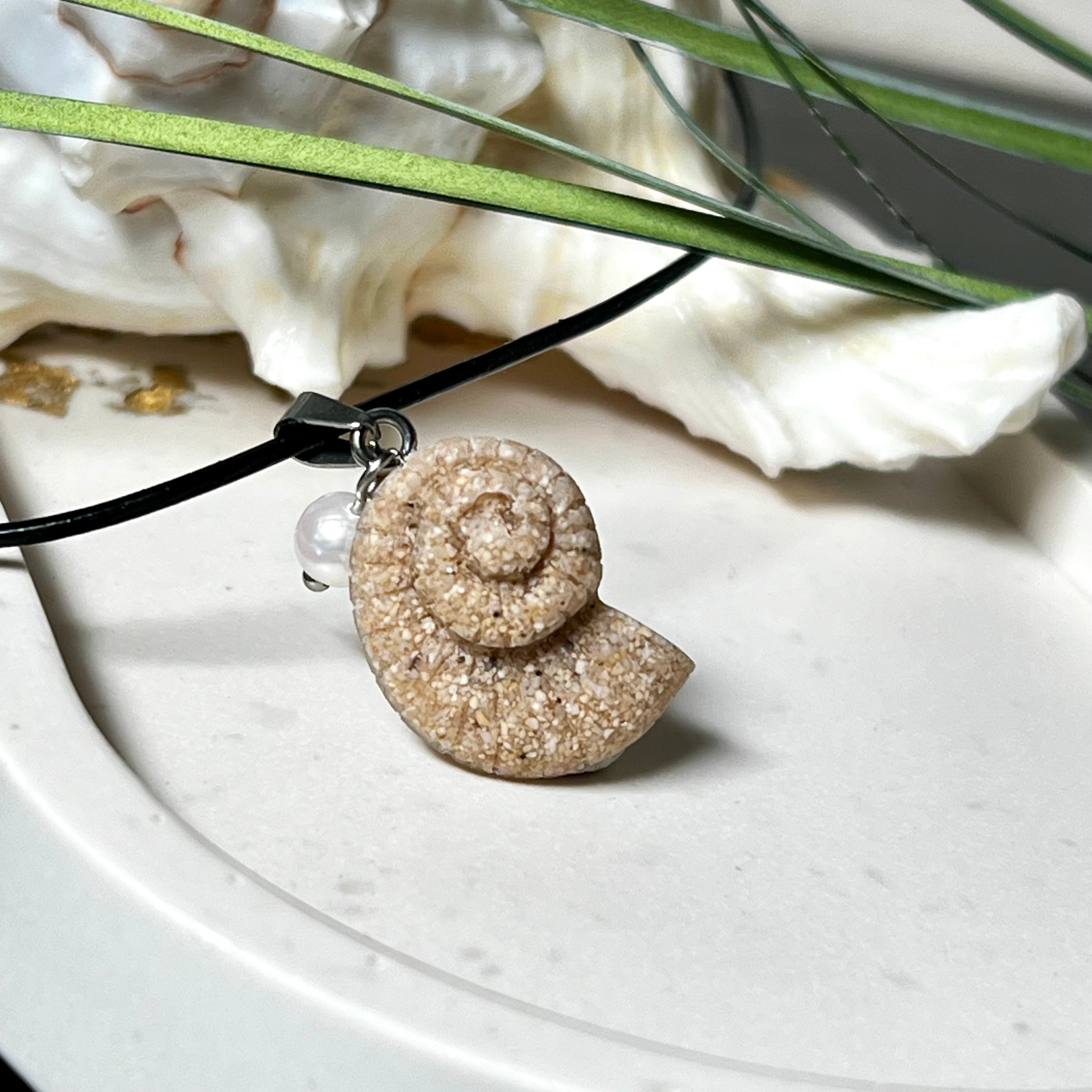 Handgefertigter Anhänger in Ammonit-Form aus echtem Meersand und Harz, kombiniert mit einer weißen Süßwasserperle. Der maritime Anhänger hängt an einer schwarzen Lederkette mit Edelstahlöse und Karabinerverschluss. Im Hintergrund liegt eine weiße Muschel, die das natürliche, strandinspirierte Design des Schmuckstücks unterstreicht. Ideal als besonderes Geschenk oder Erinnerung an den Sommer.