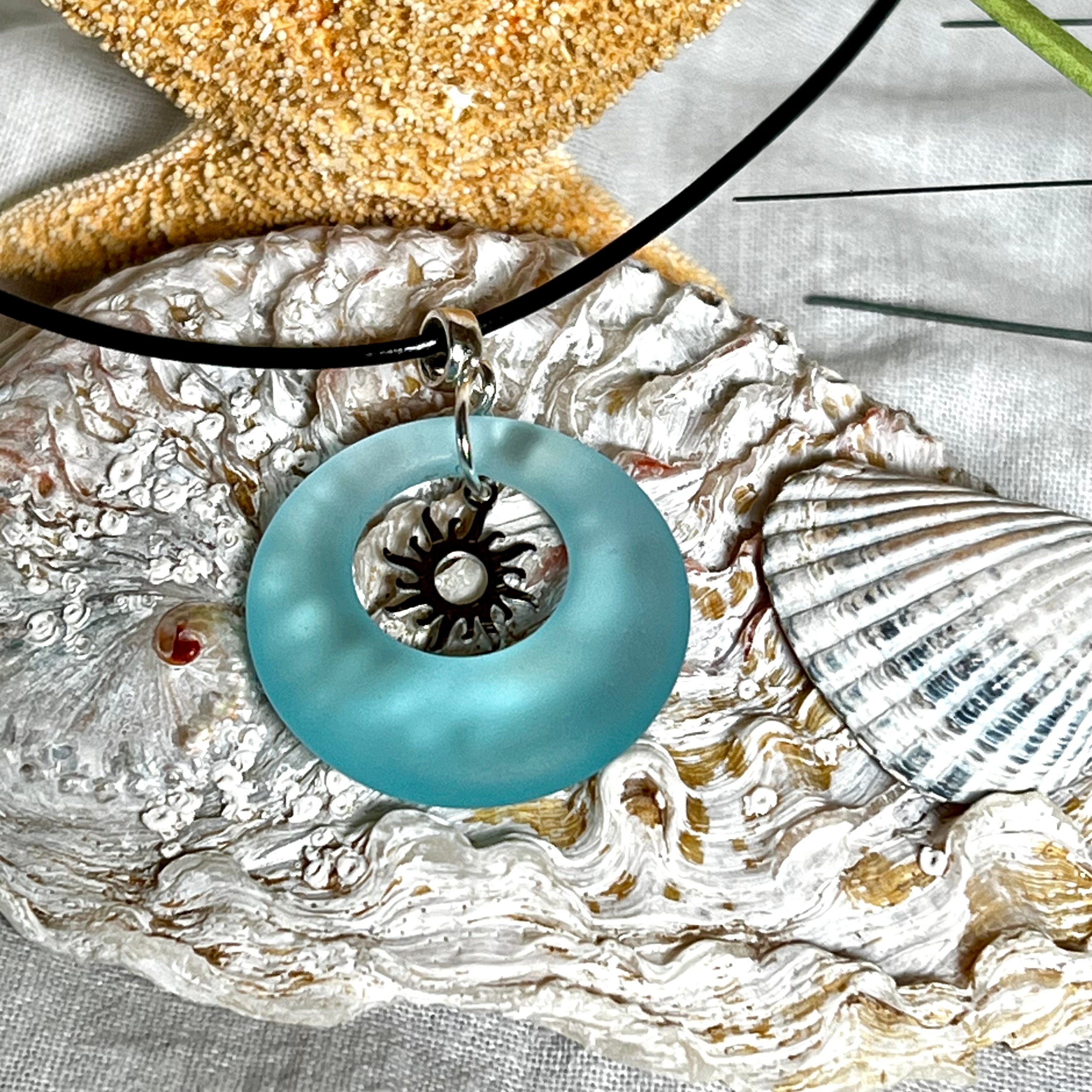Handgefertigte Halskette mit Donut-Anhänger aus türkisfarbenem Seeglas, kombiniert mit einem filigranen Sonnen-Charm aus Edelstahl. Die Kette besteht aus einer schwarzen Lederschnur (1,5 mm) mit Karabiner und Verlängerungskette aus Edelstahl. Maritimes Schmuckstück für Meerliebhaberinnen und Sommerfreunde. Unikat mit stilvoller Ausstrahlung.