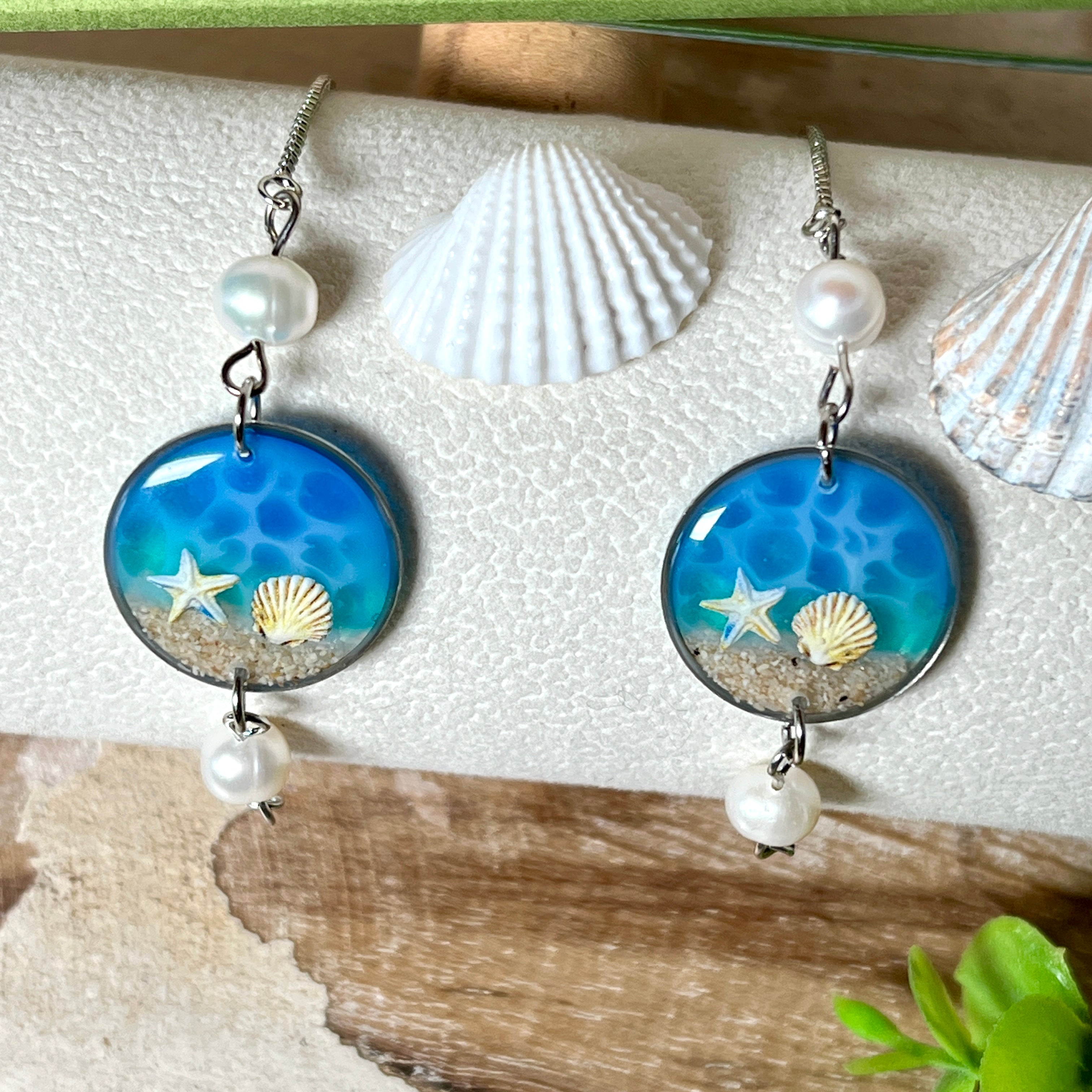Elegantes Edelstahl-Armband mit rundem Resin-Anhänger in Meeresblau. Im Anhänger liegt echter Meersand als Ufersaum; darüber Sticker-Motive Seestern & Muschel. Zwei feine Süßwasserperlen verzieren das Armband. Verstellbarer Schiebeverschluss . Handgefertigtes Unikat – maritimer Schmuck, der an Wellenrauschen und Strandspaziergänge erinnert.