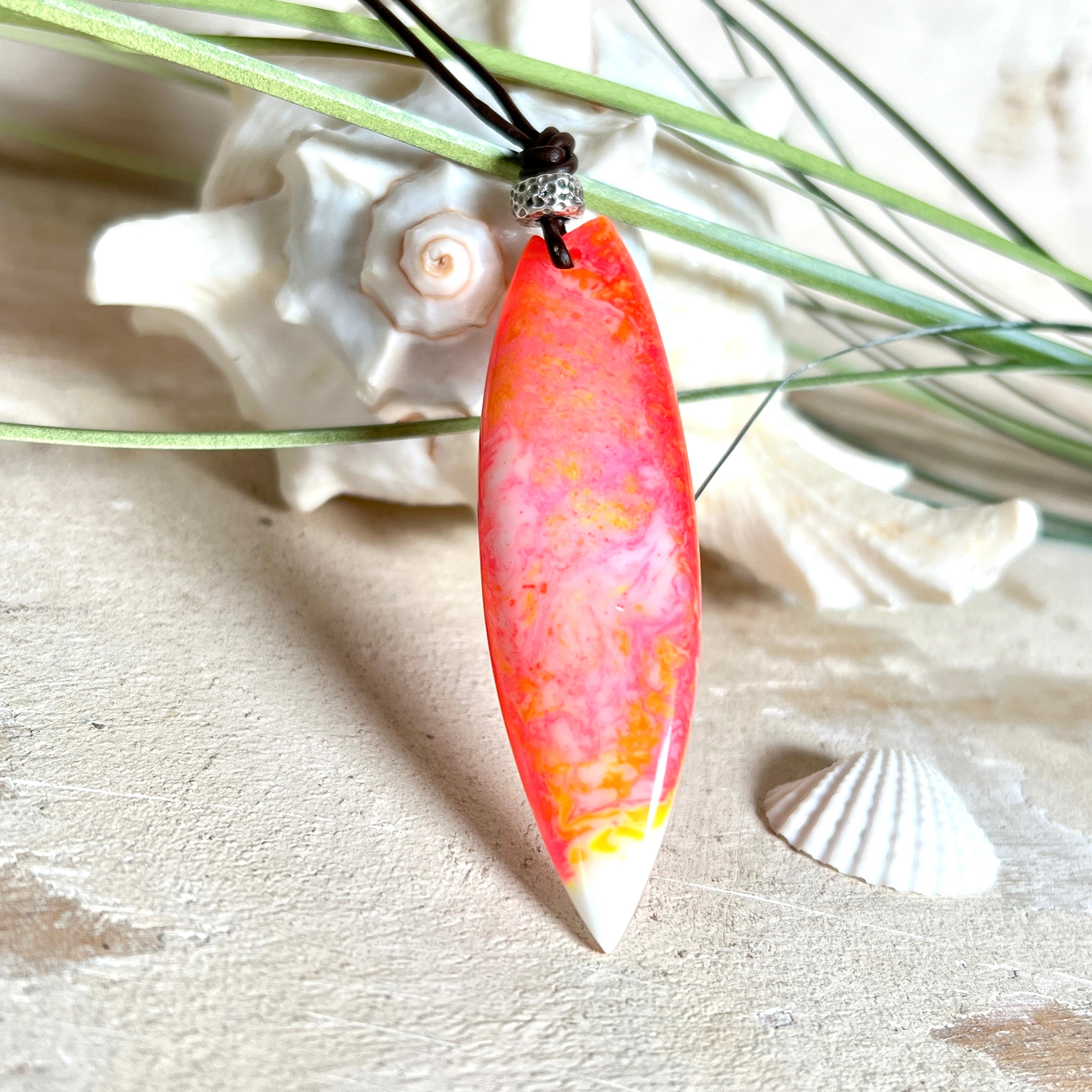 Handgefertigter Surfbrett-Anhänger aus fluoreszierendem Epoxidharz in leuchtenden Orange-, Rot- und Gelbtönen. Leuchtet bei Schwarzlicht. Kombiniert mit verstellbarem Lederband (26–45 cm), verziert mit Spacer- und Holzperlen. Das Design ist inspiriert von der Farbintensität eines Sonnenuntergangs über dem Meer.
