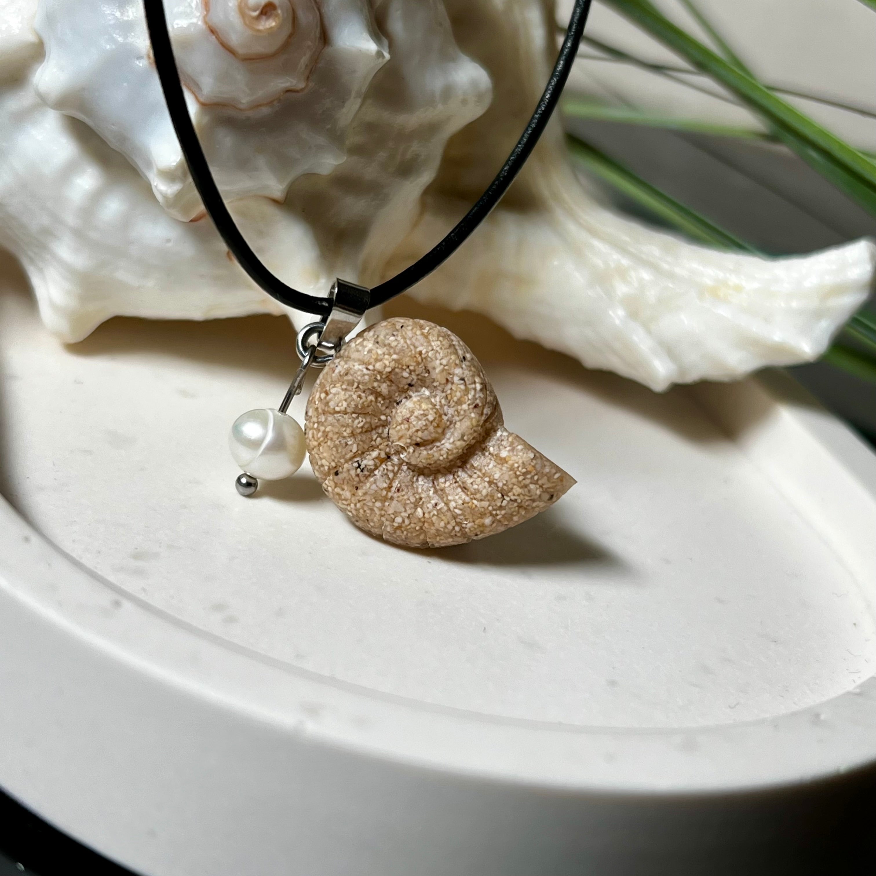 Handgefertigter Ammonit-Anhänger aus echtem Meersand & Harz mit filigraner Edelstahlöse und einer edlen Süßwasserperle. Maritimer Schmuck, inspiriert von der Kraft und Schönheit des Ozeans. Jedes Stück ein Unikat. Präsentiert auf einer Muschel, mit Lederkette.