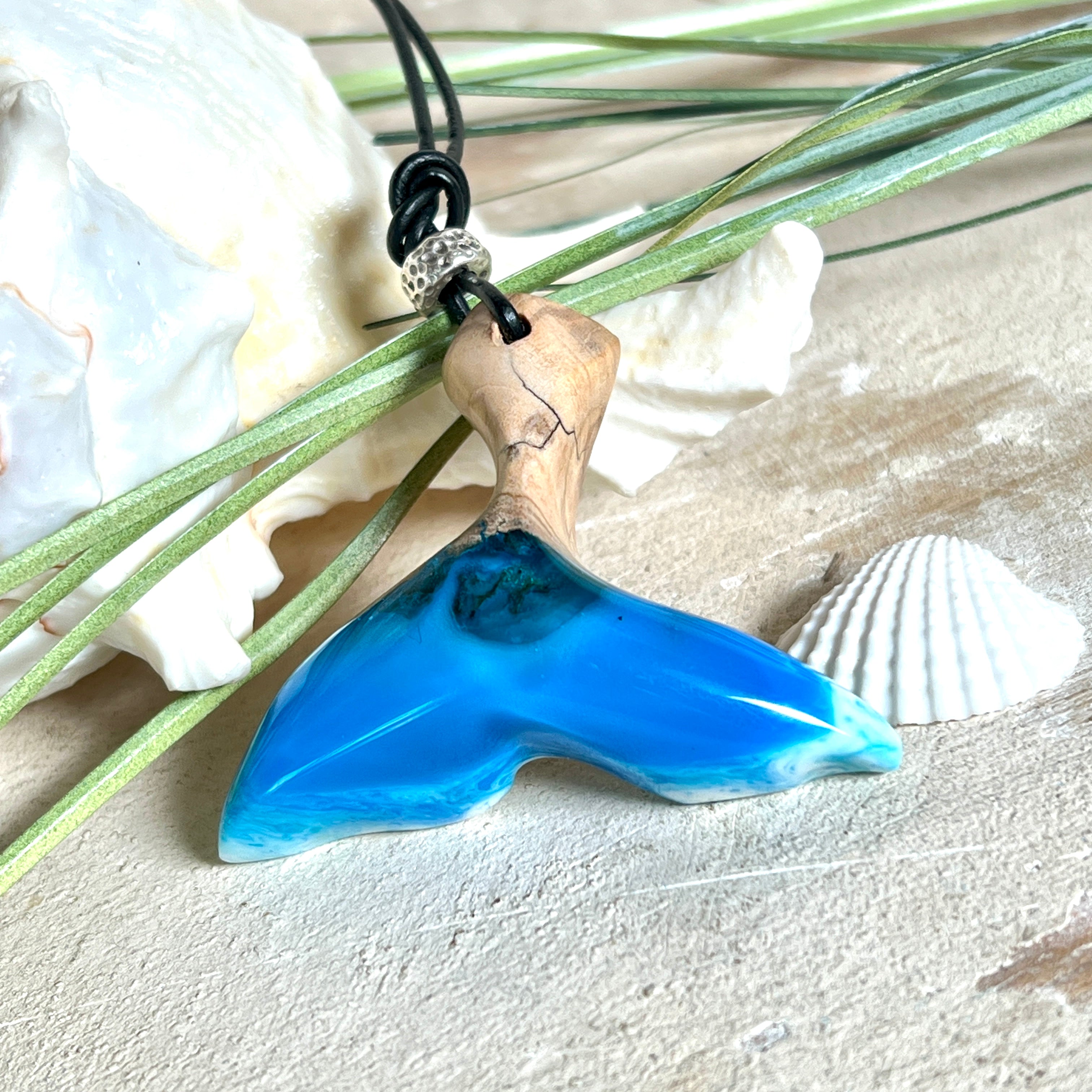 Handgefertigter Walflossen-Anhänger aus Olivenholz und glänzendem Harz in Blau-Grün-Weiß – maritimer Schmuck „Flossenklang“. Der Anhänger erinnert an die Form einer Walflosse und vereint die natürliche Maserung des Holzes mit dem schimmernden Farbspiel des Harzes. Mit verstellbarem schwarzen Lederband und Spacerperle. Ideal für Meeresliebhaber und Naturliebhaber – jedes Stück ein Unikat.