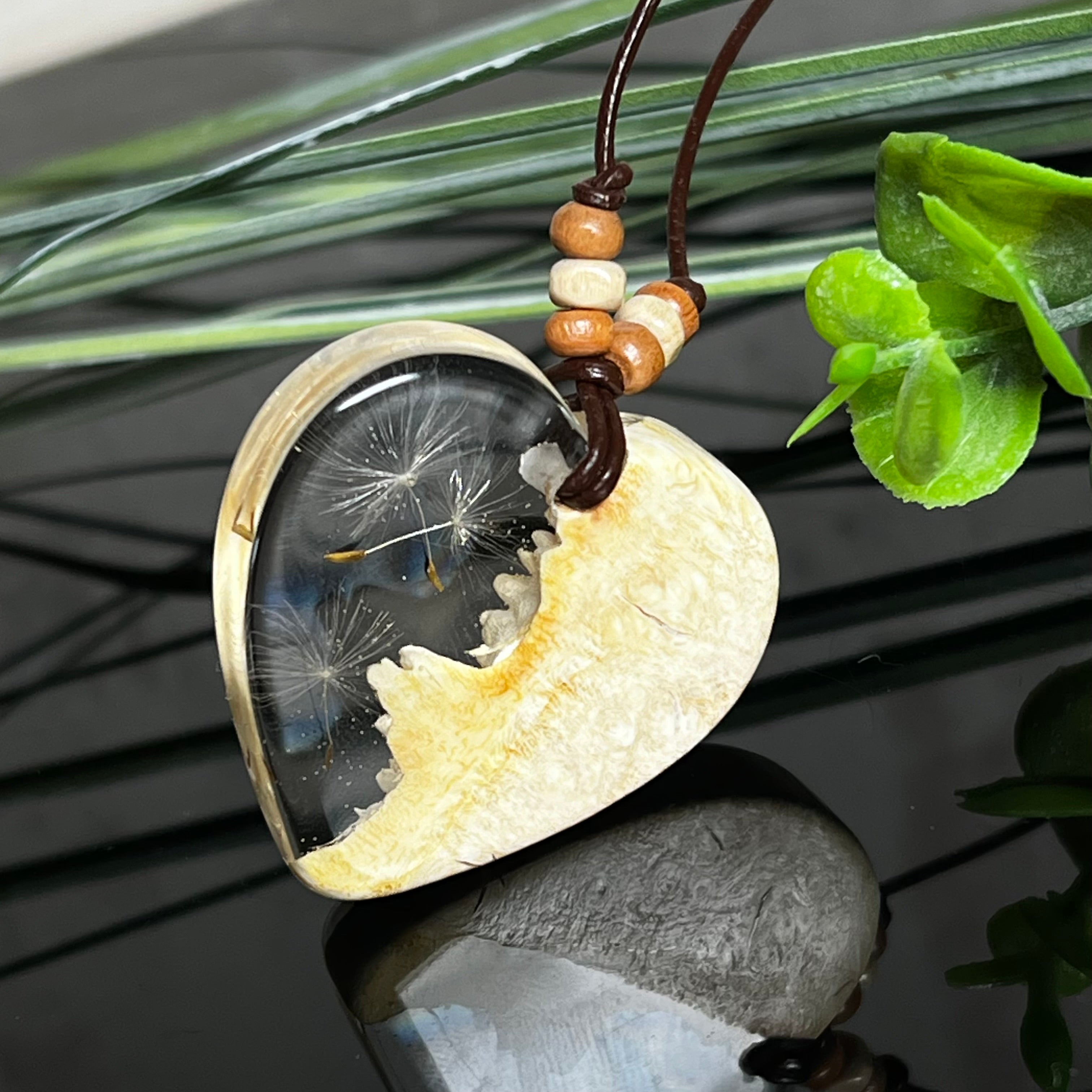 Herzanhänger aus Goldfield-Maserholz und transparentem Epoxidharz mit echter Pusteblume. Das handgefertigte Unikat kombiniert die natürliche Maserung des Holzes mit der zarten Leichtigkeit schwebender Pusteblumenschirmchen. Das braune Lederband mit Holzperlen ist verstellbar und verleiht dem Schmuckstück eine naturnahe, elegante Ausstrahlung.