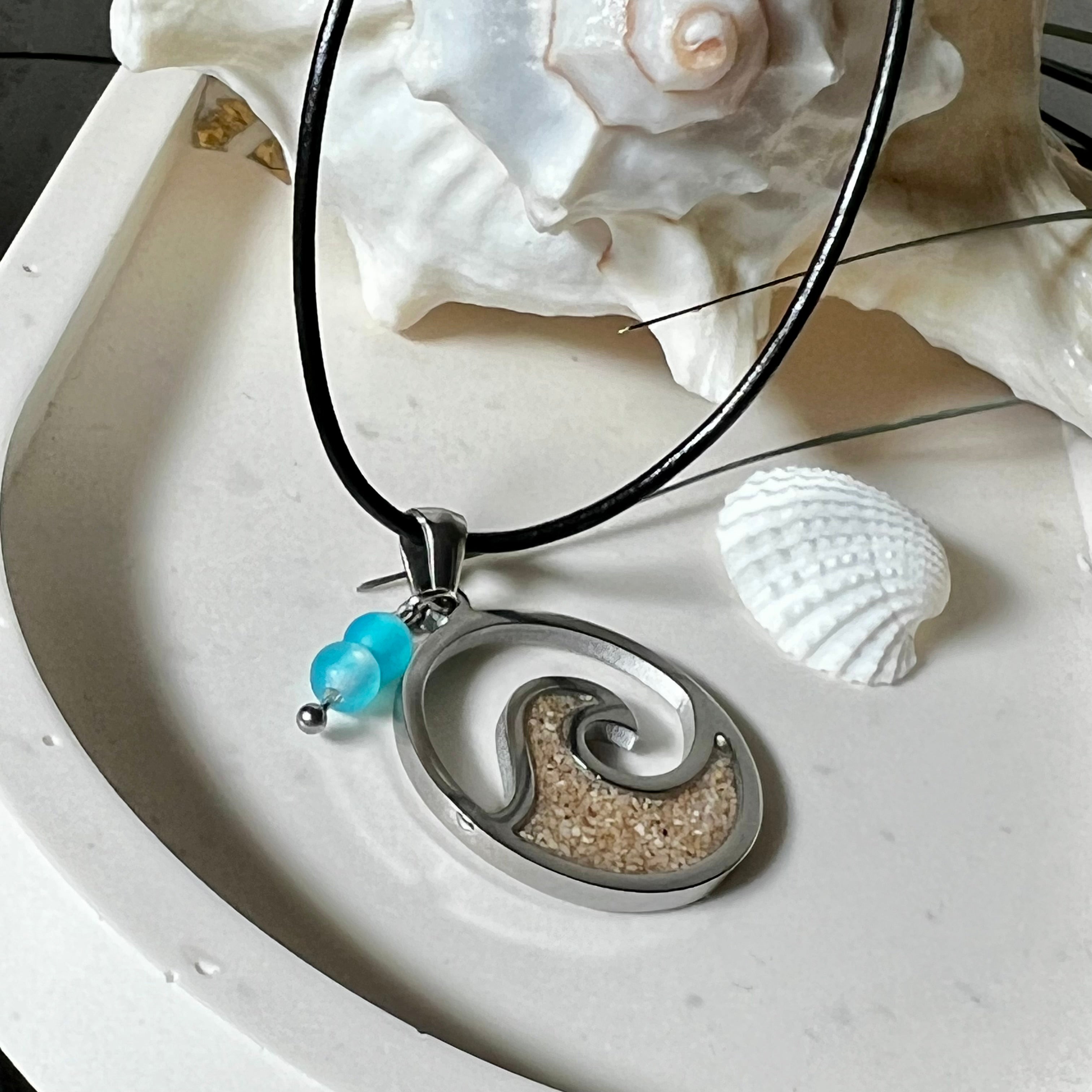 Wave Spirit – runder Edelstahlanhänger mit echter Sandfüllung in Harz eingebettet, inspiriert von einer stilisierten Welle. Verziert mit zwei blauen Achatperlen. Der Anhänger (ca. 2,5 cm Durchmesser) hängt an einer 45 cm langen Lederkette mit Edelstahlverschluss und 5 cm Verlängerungskette. Maritimer Look trifft moderne Eleganz – ein handgefertigtes Schmuckstück für Meerliebhaber