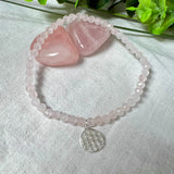 Handgefertigtes Rosenquarz-Armband mit facettierten 4mm Perlen, ergänzt durch einen filigranen Blume-des-Lebens-Anhänger aus 925er Silber. Präsentiert auf hellem Hintergrund mit Rosenquarzsteinen und goldenen Dekoelementen. Spiritueller Schmuck mit harmonisierender Wirkung.