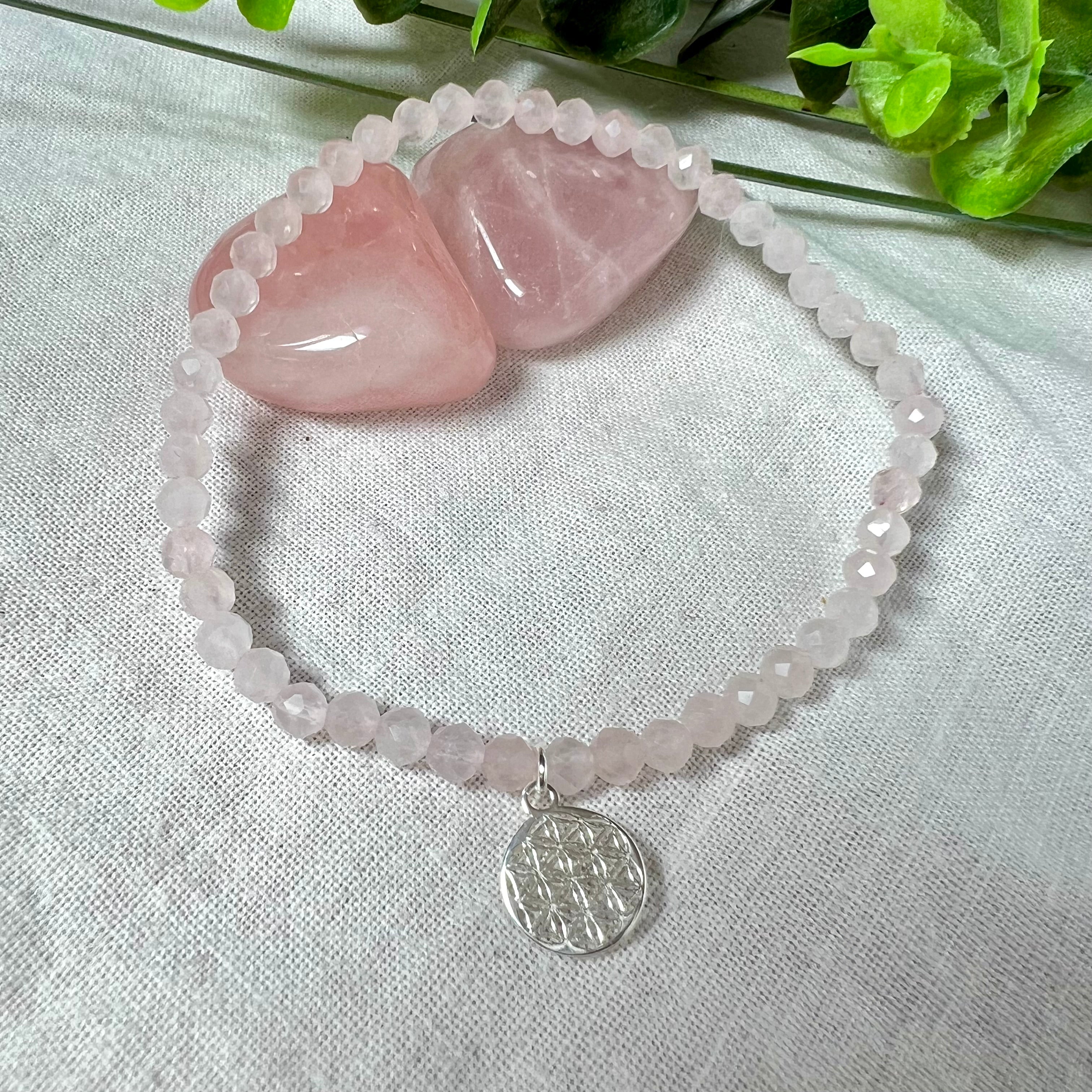 Handgefertigtes Rosenquarz-Armband mit facettierten 4mm Perlen, ergänzt durch einen filigranen Blume-des-Lebens-Anhänger aus 925er Silber. Präsentiert auf hellem Hintergrund mit Rosenquarzsteinen und goldenen Dekoelementen. Spiritueller Schmuck mit harmonisierender Wirkung.
