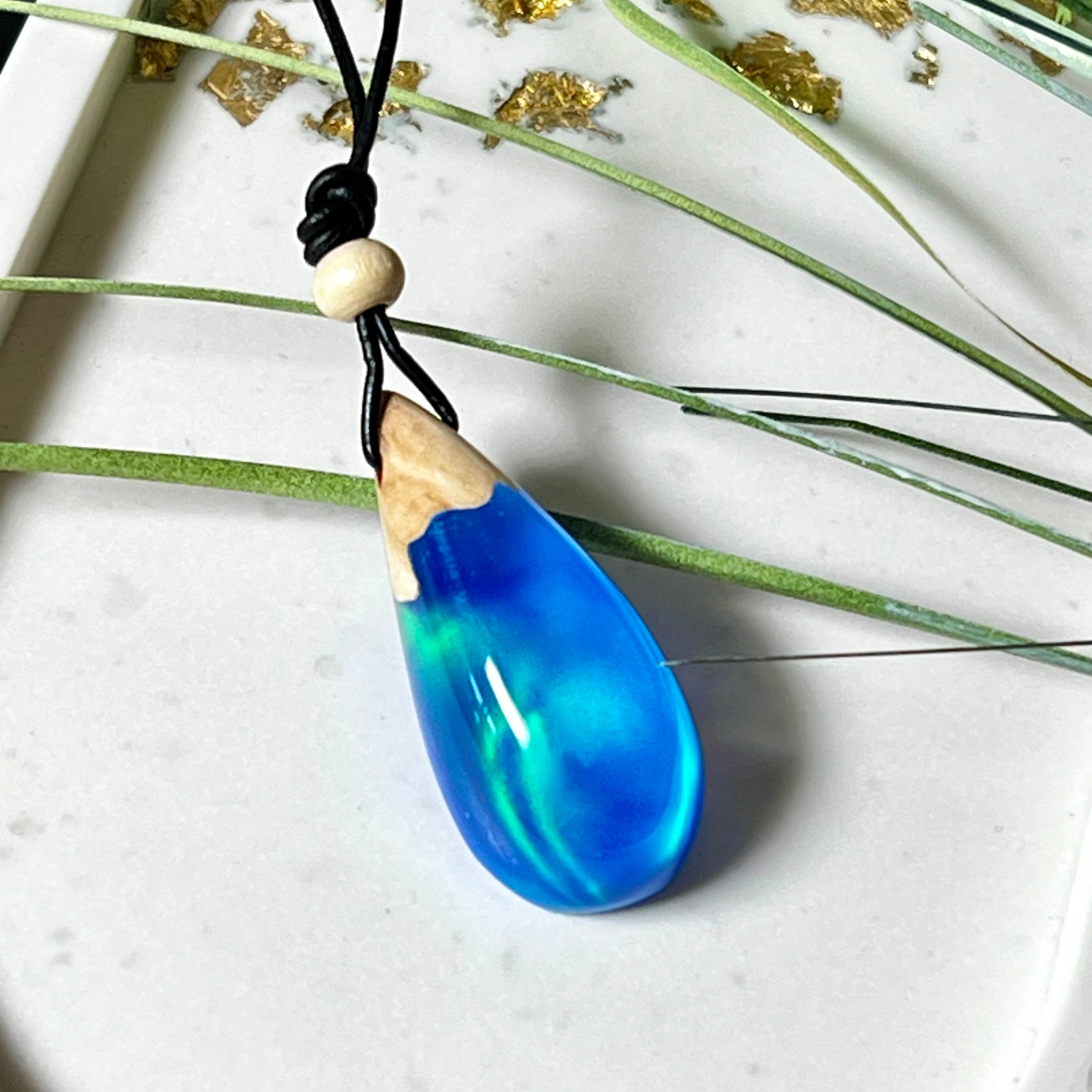 Handgefertigter Tropfenanhänger aus Maserholz und Harz mit synthetischem Opal. Die Farben schimmern je nach Lichteinfall in leuchtendem Blau und Grün – inspiriert von Polarlichtern. Der Anhänger ist ca. 3,9 × 1,8 cm groß und hängt an einem verstellbaren Lederband mit Holzperlen. Jedes Stück ist ein Unikat mit einzigartiger Holzmaserung. Ideal als Geschenk oder stilvoller Begleiter im Alltag.