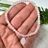 Handgefertigtes Rosenquarz-Armband mit facettierten 4mm Perlen, ergänzt durch einen filigranen Blume-des-Lebens-Anhänger aus 925er Silber. Präsentiert auf hellem Hintergrund mit Rosenquarzsteinen und goldenen Dekoelementen. Spiritueller Schmuck mit harmonisierender Wirkung.
