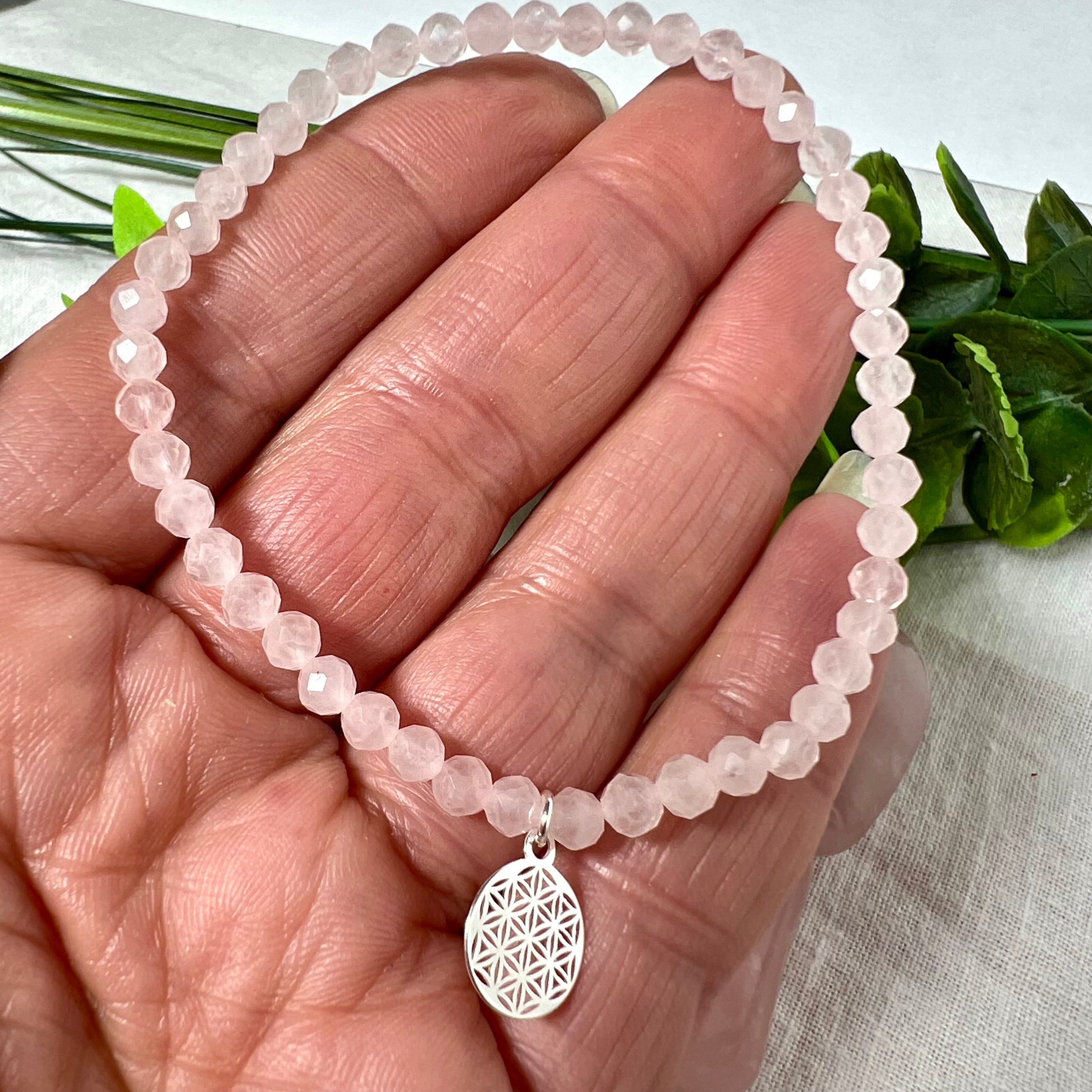 Handgefertigtes Rosenquarz-Armband mit facettierten 4mm Perlen, ergänzt durch einen filigranen Blume-des-Lebens-Anhänger aus 925er Silber. Präsentiert auf hellem Hintergrund mit Rosenquarzsteinen und goldenen Dekoelementen. Spiritueller Schmuck mit harmonisierender Wirkung.