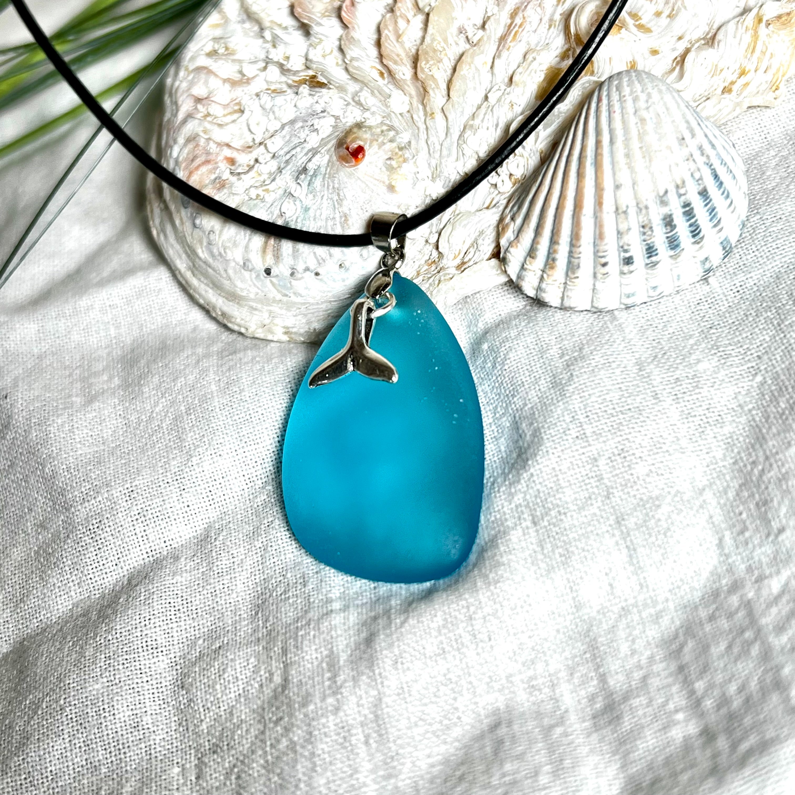 Edle Halskette „Blue Essence“ mit handgefertigtem türkisfarbenem Seeglas-Anhänger und Edelstahl-Flosse. Die Kette besteht aus schwarzer Lederkordel mit Karabinerverschluss und Verlängerung aus Edelstahl. Das matte Seeglas reflektiert Licht wie Meereswasser – ideal für Strandliebhaber und Naturliebhaber.
