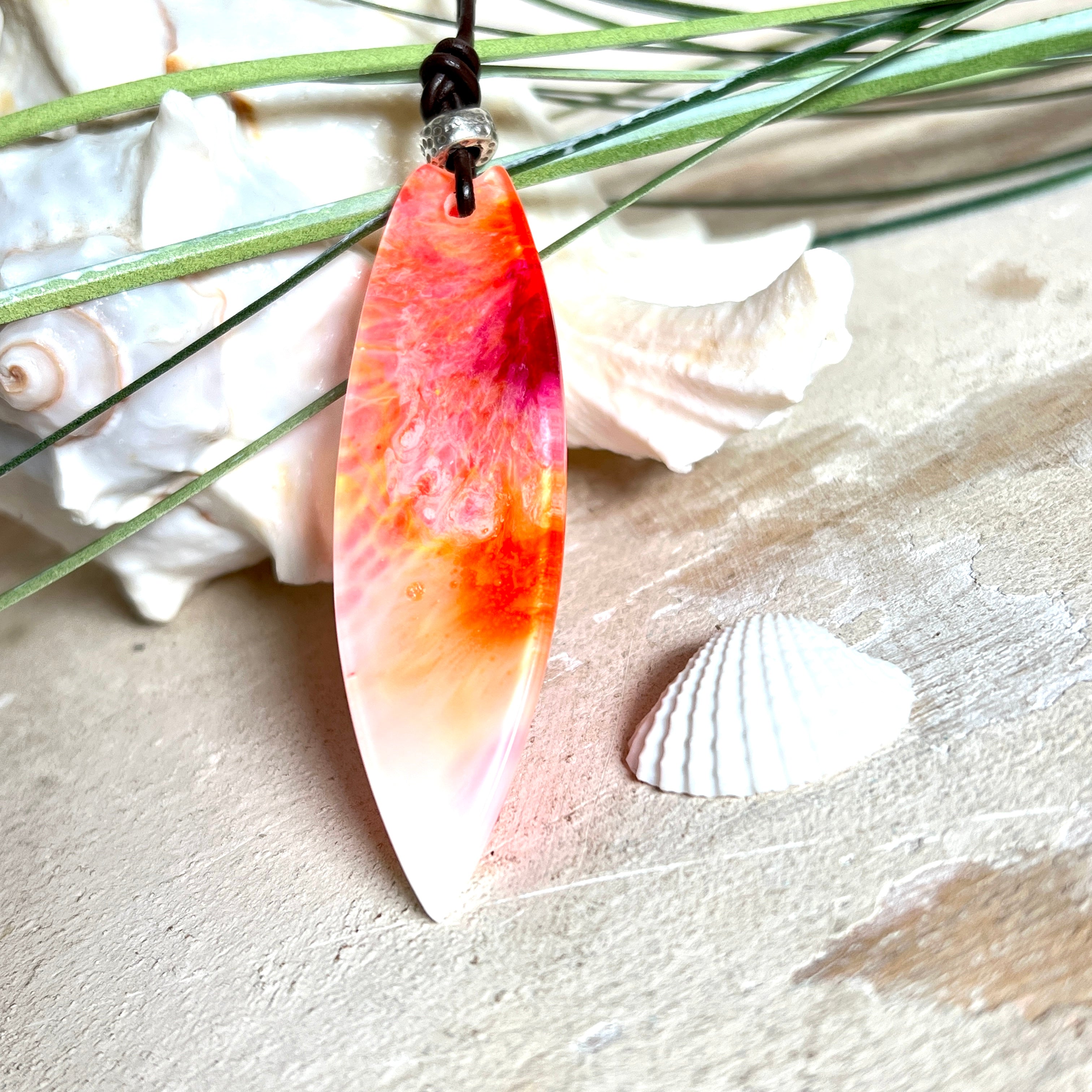 Handgefertigter Surfbrett-Anhänger aus fluoreszierendem Epoxidharz in leuchtenden Orange-, Rot- und Gelbtönen. Leuchtet bei Schwarzlicht. Kombiniert mit verstellbarem Lederband (26–45 cm), verziert mit Spacer- und Holzperlen. Das Design ist inspiriert von der Farbintensität eines Sonnenuntergangs über dem Meer.