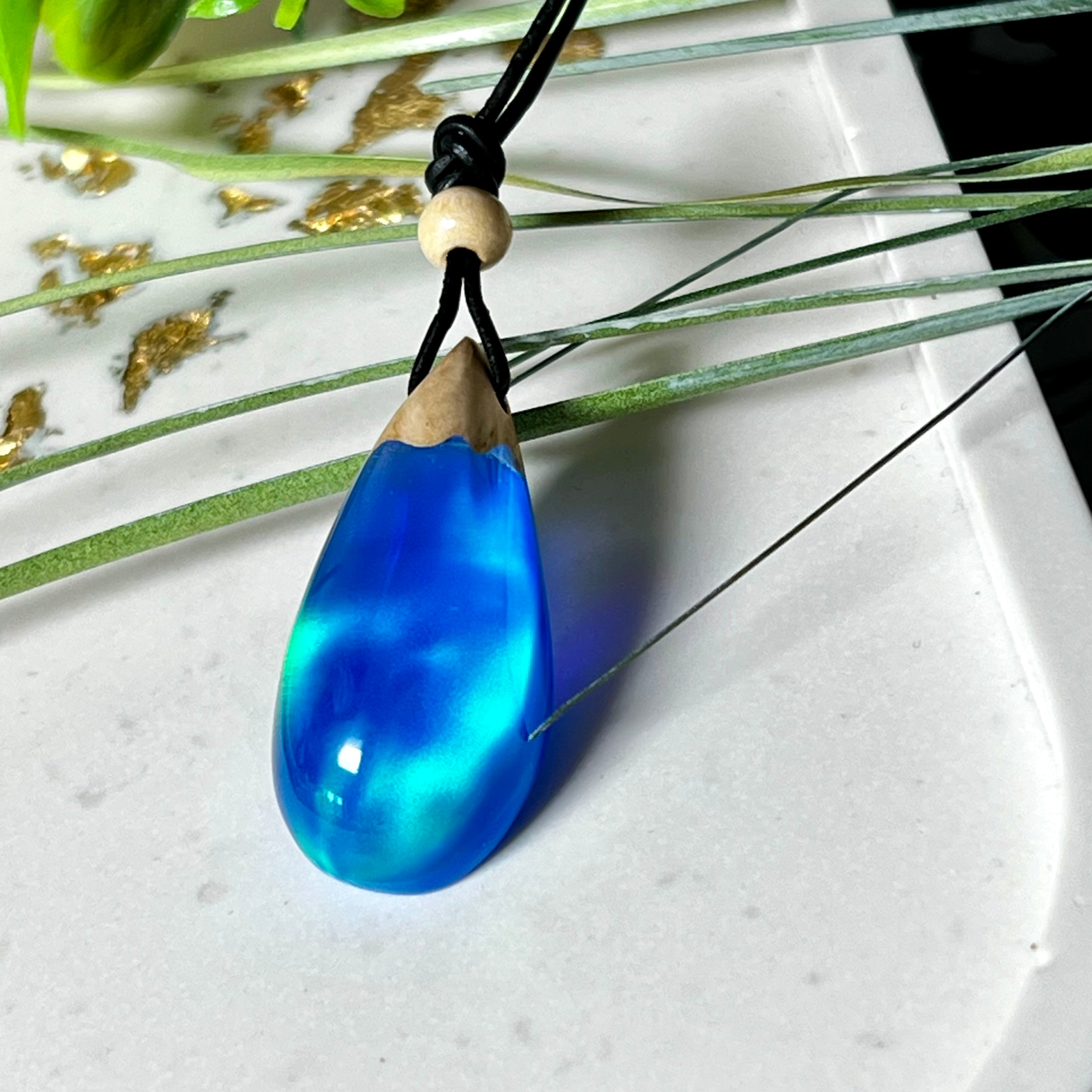 Handgefertigter Tropfenanhänger aus Maserholz und Harz mit synthetischem Opal. Die Farben schimmern je nach Lichteinfall in leuchtendem Blau und Grün – inspiriert von Polarlichtern. Der Anhänger ist ca. 3,9 × 1,8 cm groß und hängt an einem verstellbaren Lederband mit Holzperlen. Jedes Stück ist ein Unikat mit einzigartiger Holzmaserung. Ideal als Geschenk oder stilvoller Begleiter im Alltag.