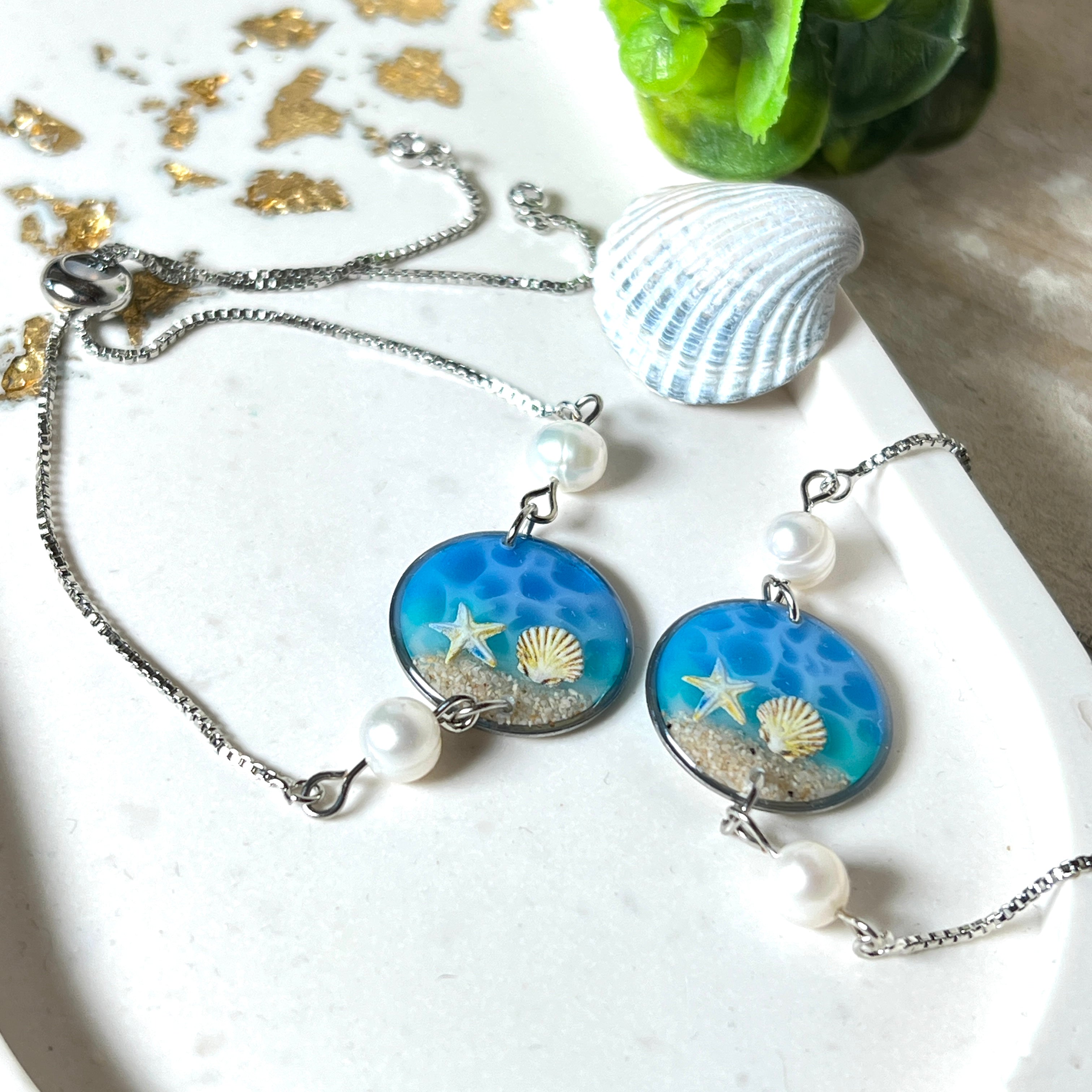 Elegantes Edelstahl-Armband mit rundem Resin-Anhänger in Meeresblau. Im Anhänger liegt echter Meersand als Ufersaum; darüber Sticker-Motive Seestern & Muschel. Zwei feine Süßwasserperlen verzieren das Armband. Verstellbarer Schiebeverschluss . Handgefertigtes Unikat – maritimer Schmuck, der an Wellenrauschen und Strandspaziergänge erinnert.