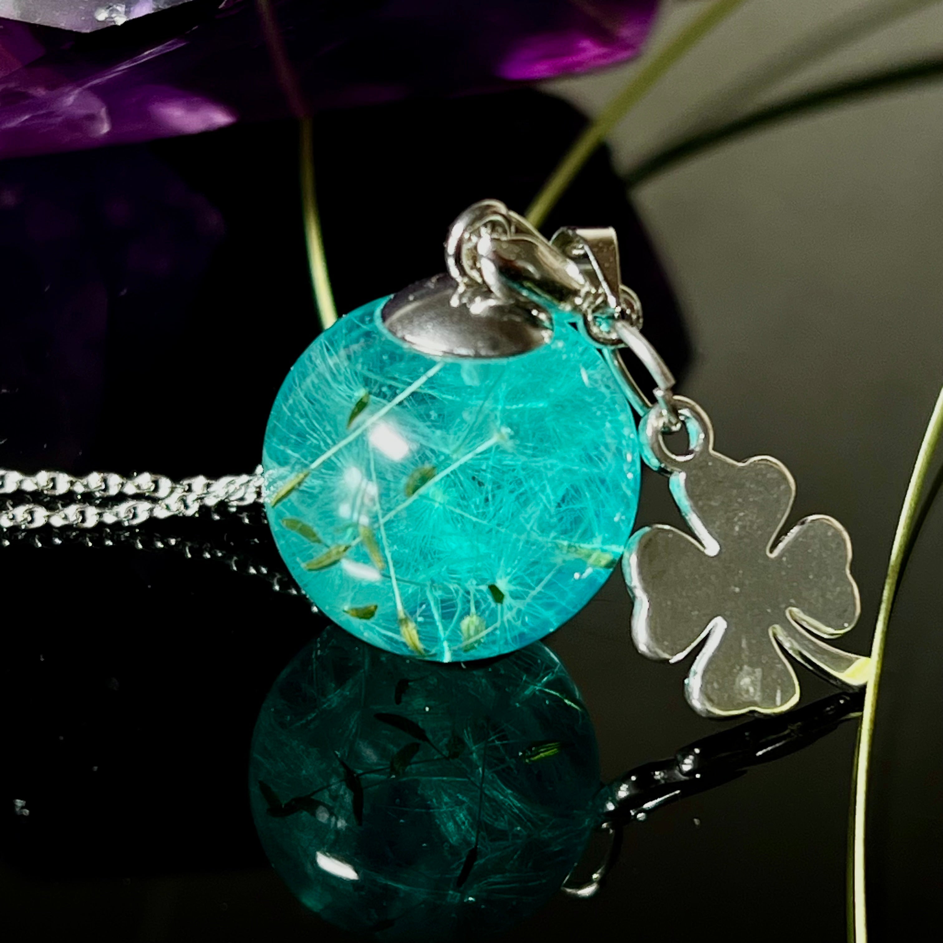 Eine handgefertigten Halskette mit türkiser Harzkugel, in der echte Pusteblumensamen eingeschlossen sind. Ergänzt durch einen glänzenden Edelstahl-Kleeblatt-Charm und einer feinen rhodinierten 925 Silberkette. Der Anhänger symbolisiert Glück, Leichtigkeit und neue Wege – ein emotionales Schmuckstück mit Bedeutung.