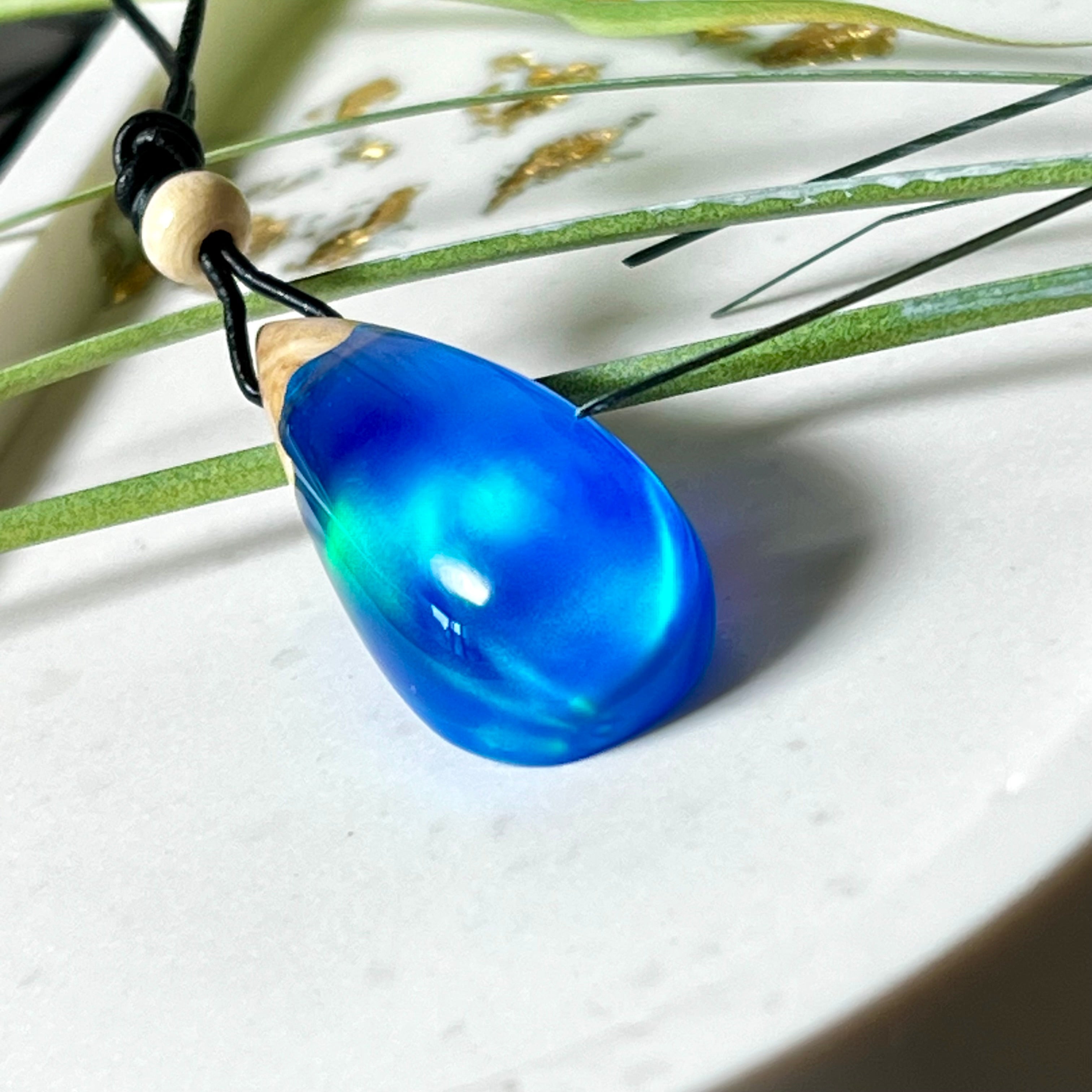 Handgefertigter Tropfenanhänger aus Maserholz und Harz mit synthetischem Opal. Die Farben schimmern je nach Lichteinfall in leuchtendem Blau und Grün – inspiriert von Polarlichtern. Der Anhänger ist ca. 3,9 × 1,8 cm groß und hängt an einem verstellbaren Lederband mit Holzperlen. Jedes Stück ist ein Unikat mit einzigartiger Holzmaserung. Ideal als Geschenk oder stilvoller Begleiter im Alltag.