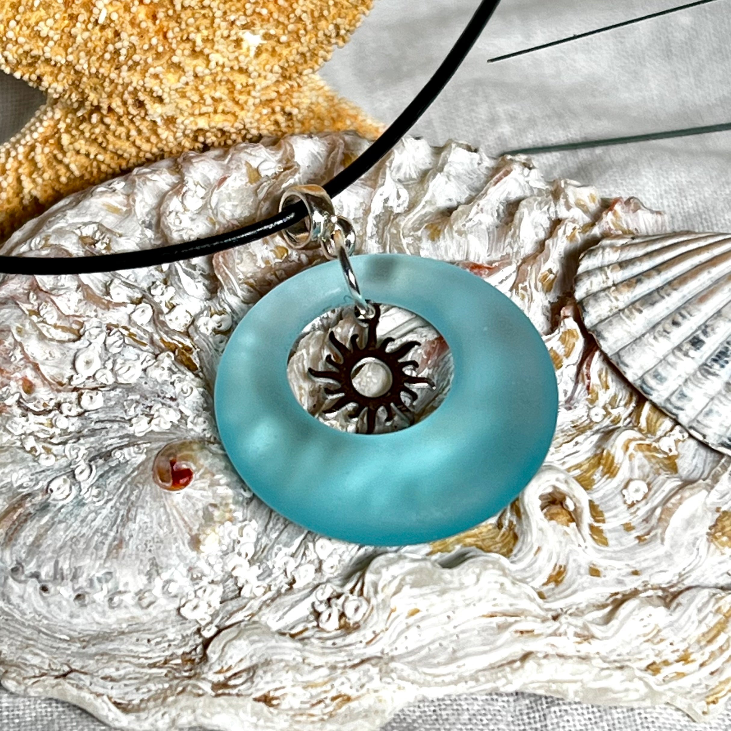 Handgefertigte Halskette mit Donut-Anhänger aus türkisfarbenem Seeglas, kombiniert mit einem filigranen Sonnen-Charm aus Edelstahl. Die Kette besteht aus einer schwarzen Lederschnur (1,5 mm) mit Karabiner und Verlängerungskette aus Edelstahl. Maritimes Schmuckstück für Meerliebhaberinnen und Sommerfreunde. Unikat mit stilvoller Ausstrahlung.