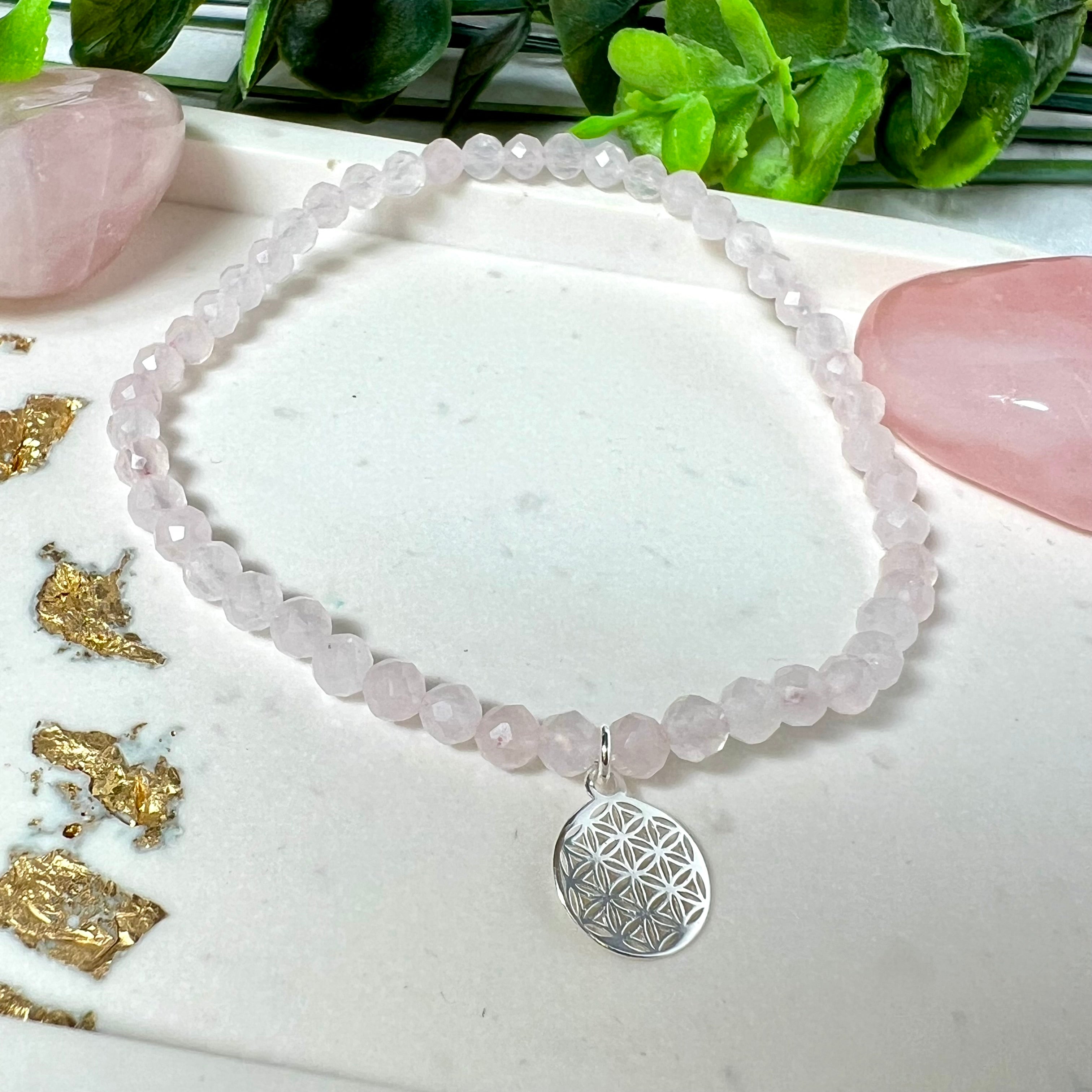 Handgefertigtes Rosenquarz-Armband mit facettierten 4mm Perlen, ergänzt durch einen filigranen Blume-des-Lebens-Anhänger aus 925er Silber. Präsentiert auf hellem Hintergrund mit Rosenquarzsteinen und goldenen Dekoelementen. Spiritueller Schmuck mit harmonisierender Wirkung.