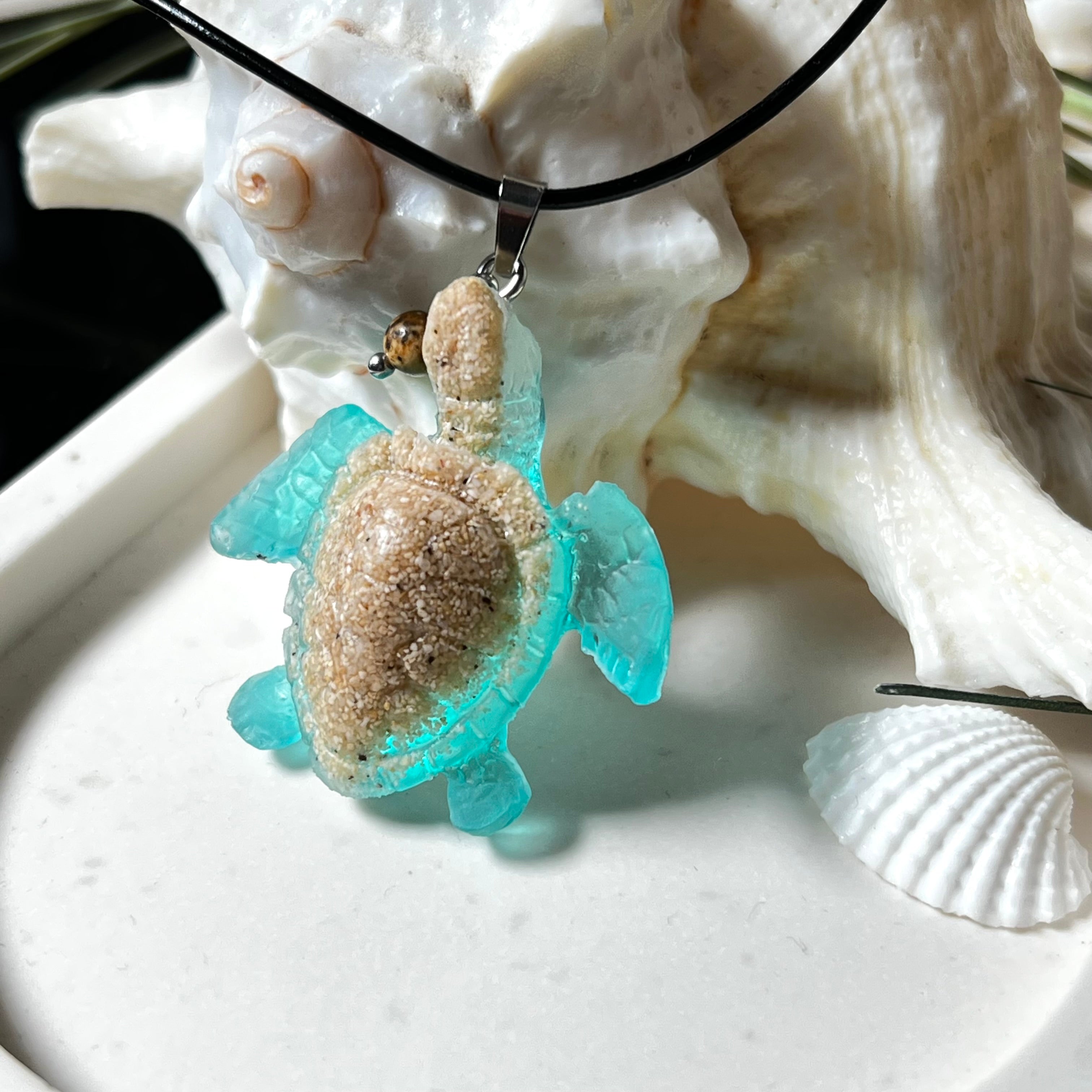 Handgefertigter Schildkröten-Anhänger Turtle Harmony aus echtem Meersand und transparent-türkisfarbenem Harz, kombiniert mit einer kleinen Jaspisperle. Symbol für Weisheit, Schutz und Harmonie. Mit Edelstahl-Anhängerschlaufe und Lederkette (45 + 5 cm). Maritimer Schmuck als Unikat, ideal als liebevolles Geschenk oder stilvolles Accessoire für den Alltag.