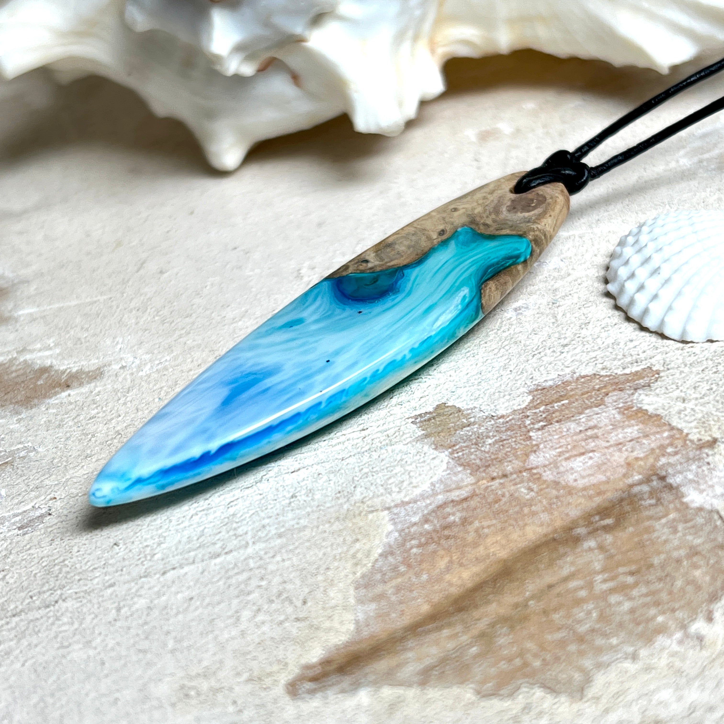 Surfbrett-Anhänger handgefertigt aus Maserholz & Harz in Blau-Weiß. Größe ca. 6,3 × 2 cm. Lederband verstellbar 26–47 cm, Schiebeknoten mit Holzperlen.