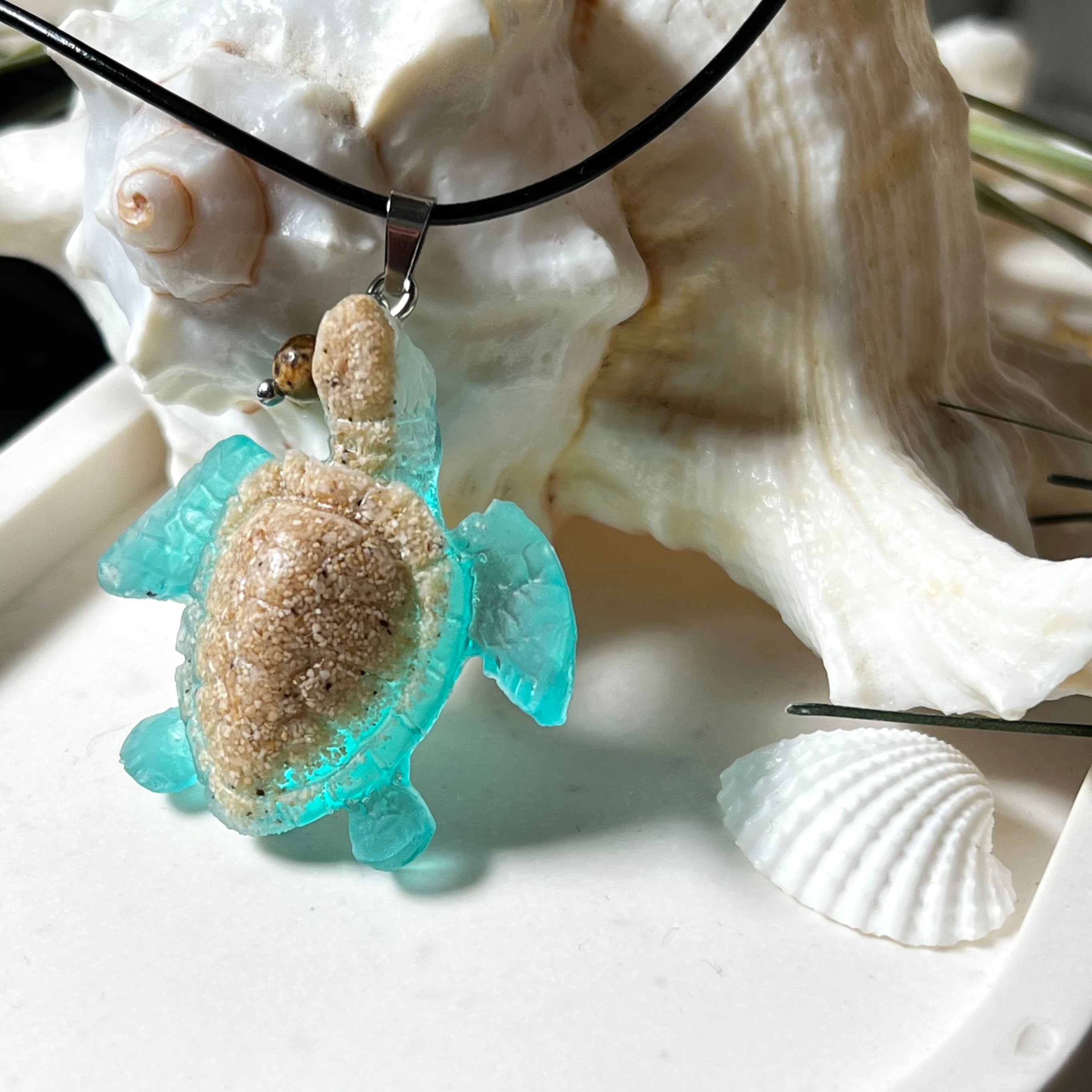Handgefertigter Schildkröten-Anhänger Turtle Harmony aus echtem Meersand und transparent-türkisfarbenem Harz, kombiniert mit einer kleinen Jaspisperle. Symbol für Weisheit, Schutz und Harmonie. Mit Edelstahl-Anhängerschlaufe und Lederkette (45 + 5 cm). Maritimer Schmuck als Unikat, ideal als liebevolles Geschenk oder stilvolles Accessoire für den Alltag.