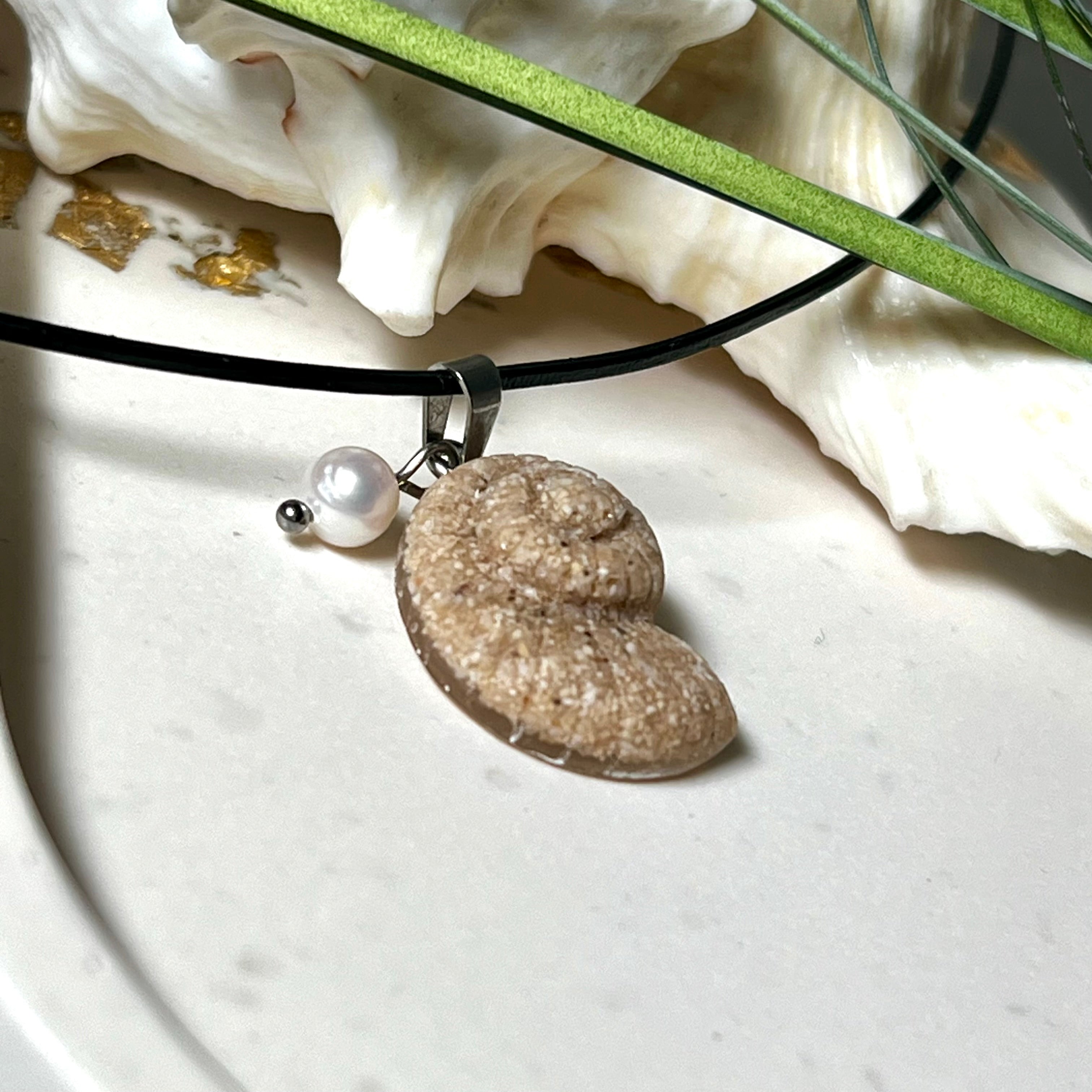 Handgefertigter Anhänger in Ammonit-Form aus echtem Meersand und Harz, kombiniert mit einer weißen Süßwasserperle. Der maritime Anhänger hängt an einer schwarzen Lederkette mit Edelstahlöse und Karabinerverschluss. Im Hintergrund liegt eine weiße Muschel, die das natürliche, strandinspirierte Design des Schmuckstücks unterstreicht. Ideal als besonderes Geschenk oder Erinnerung an den Sommer.