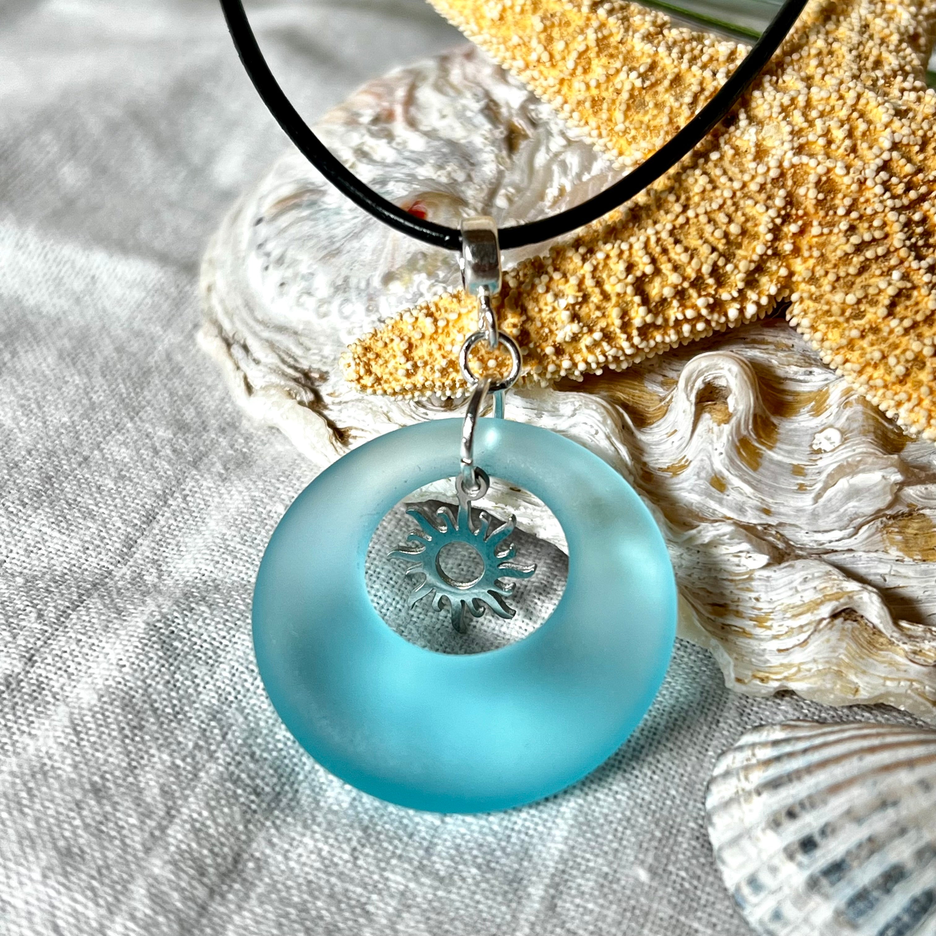 Handgefertigte Halskette mit Donut-Anhänger aus türkisfarbenem Seeglas, kombiniert mit einem filigranen Sonnen-Charm aus Edelstahl. Die Kette besteht aus einer schwarzen Lederschnur (1,5 mm) mit Karabiner und Verlängerungskette aus Edelstahl. Maritimes Schmuckstück für Meerliebhaberinnen und Sommerfreunde. Unikat mit stilvoller Ausstrahlung.