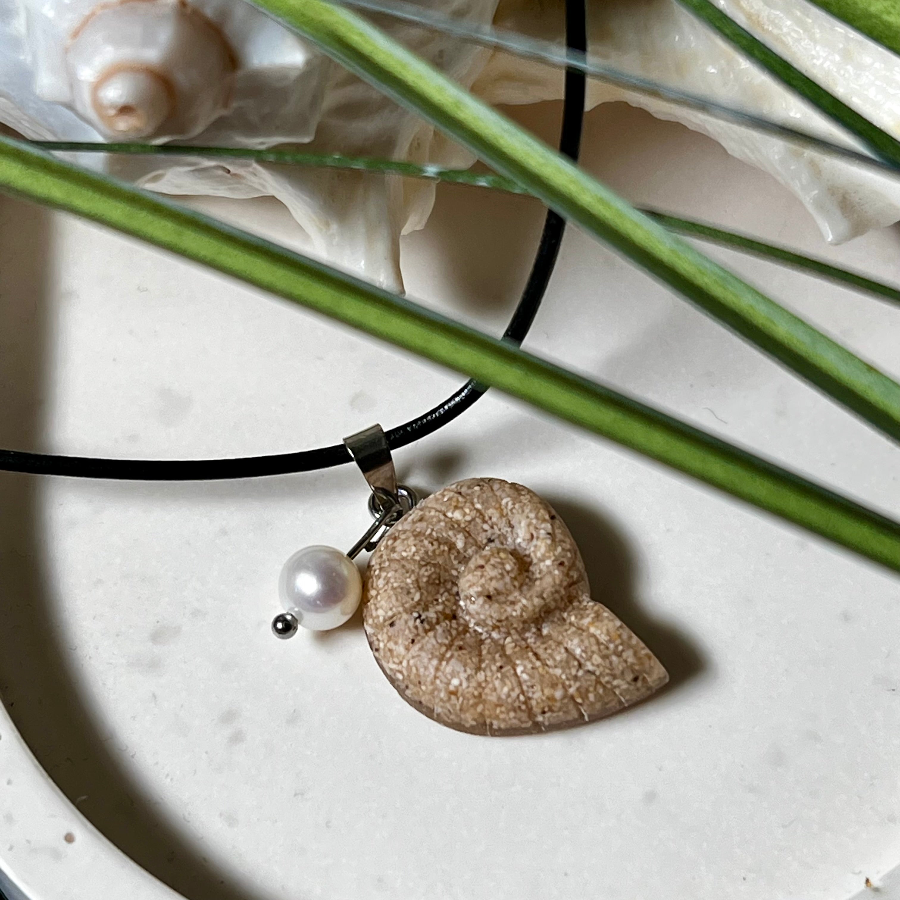 Handgefertigter Ammonit-Anhänger aus echtem Meersand & Harz mit filigraner Edelstahlöse und einer edlen Süßwasserperle. Maritimer Schmuck, inspiriert von der Kraft und Schönheit des Ozeans. Jedes Stück ein Unikat. Präsentiert auf einer Muschel, mit Lederkette.