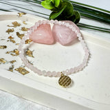„Ein filigranes Armband aus zarten Rosenquarz-Perlen mit einem goldenen Anhänger, der das Muster der Blume des Lebens darstellt. Das Armband liegt auf einem hellen Untergrund, umgeben von Rosenquarz-Steinen und goldenen Blattakzenten, mit grünen Blättern im Hintergrund.“
