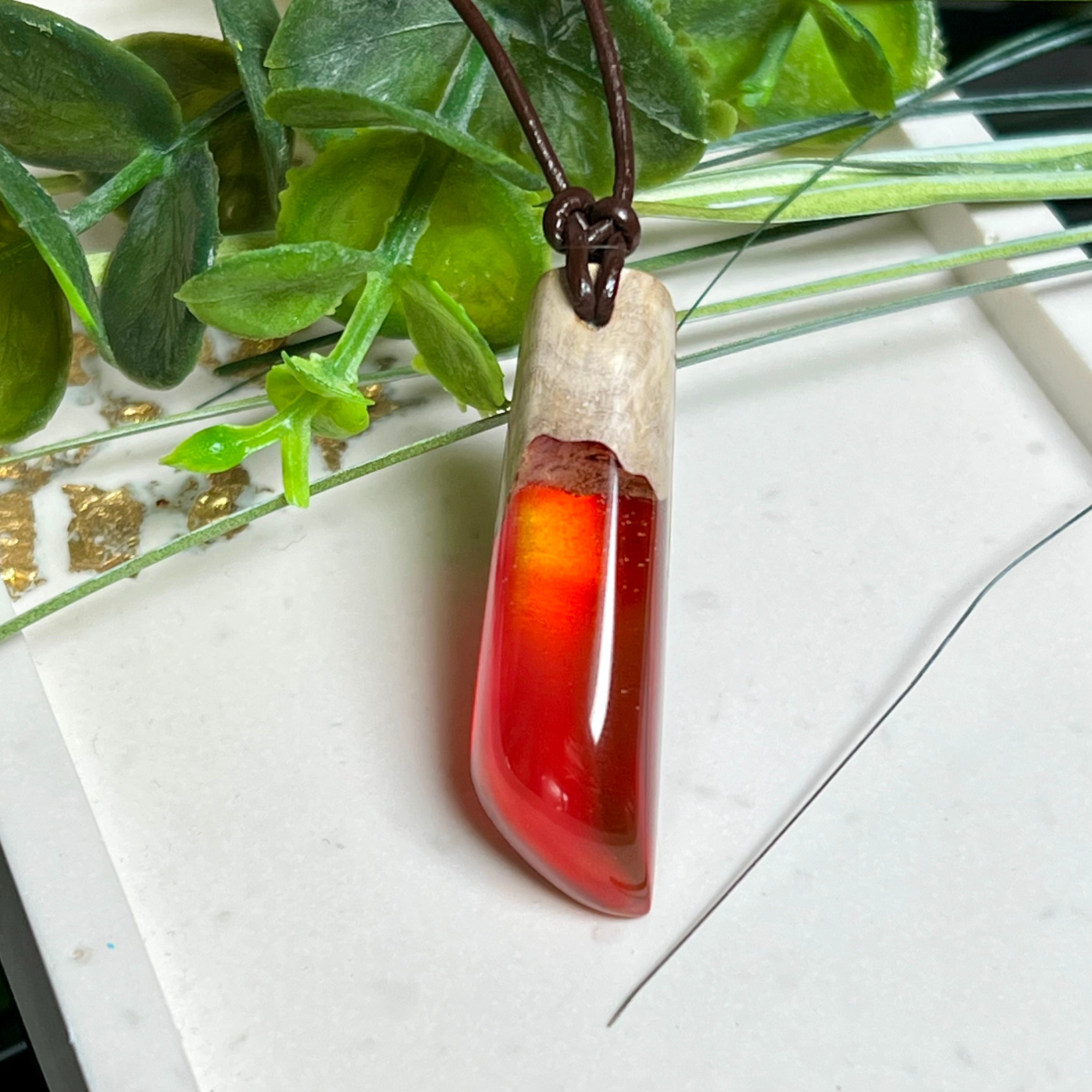 Handgefertigter Hei Toki Anhänger aus Goldfield-Maserholz und rubinrotem Labor-Opal, eingebettet in glänzendes Epoxidharz. Je nach Lichteinfall schimmert das Harz in warmen Farben mit Regenbogeneffekt. Der Anhänger ist ca. 4,3 × 1,5 cm groß und wird an einem verstellbaren Lederband mit Holzperlen getragen. Symbolischer Unikatschmuck – ideal als Geschenk für besondere Menschen.
