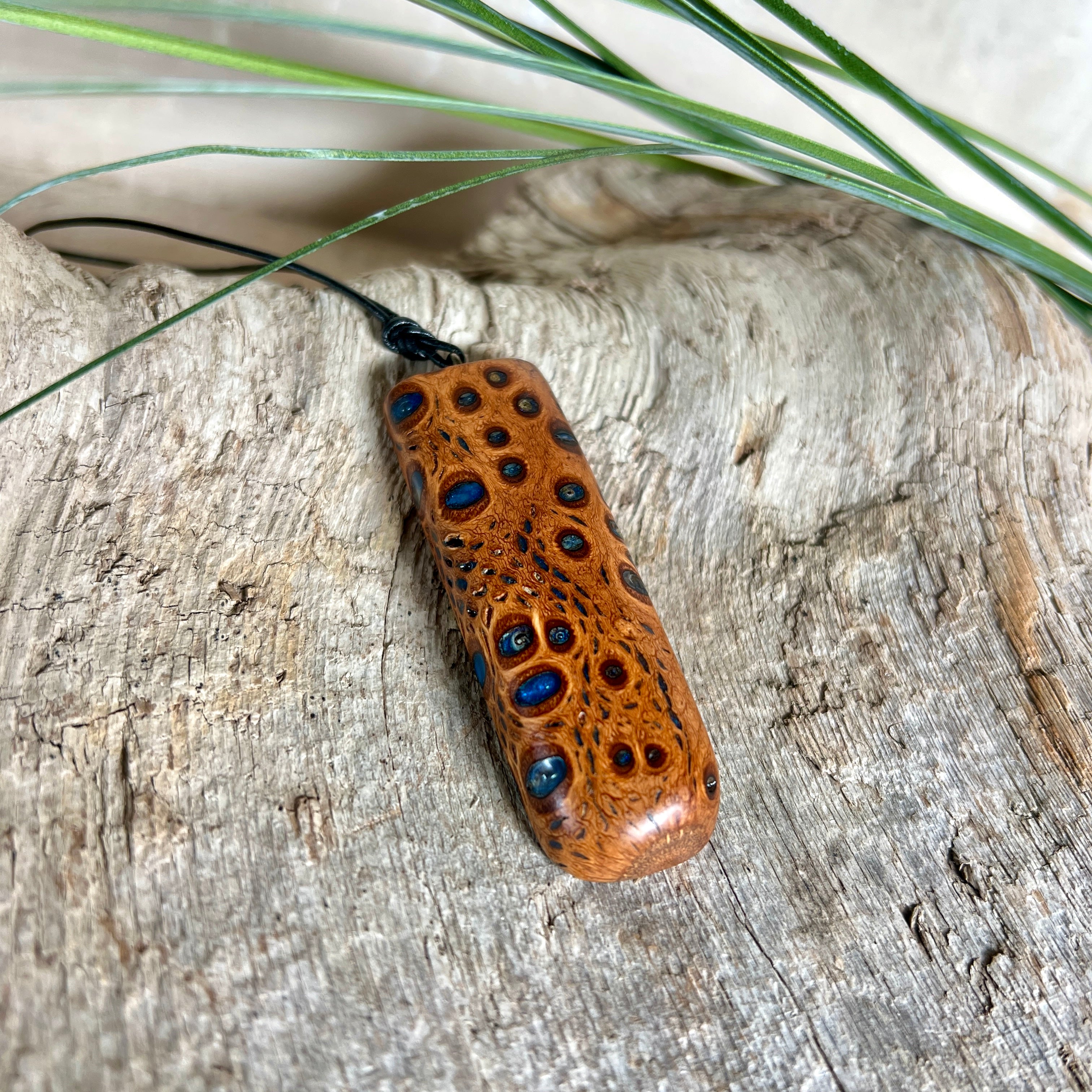 Handgefertigter Anhänger aus australischem Banksiazapfen und gegossenem Harz in Blau. Der rechteckige Anhänger (5,5 x 2 cm) zeigt eine charakteristische Maserung mit markanten Löchern, die durch die Samenstruktur des Zapfens entstehen. Das Lederband ist verstellbar (29–51 cm) und mit Holzperlen verziert. Ein edles Natur-Unikat.