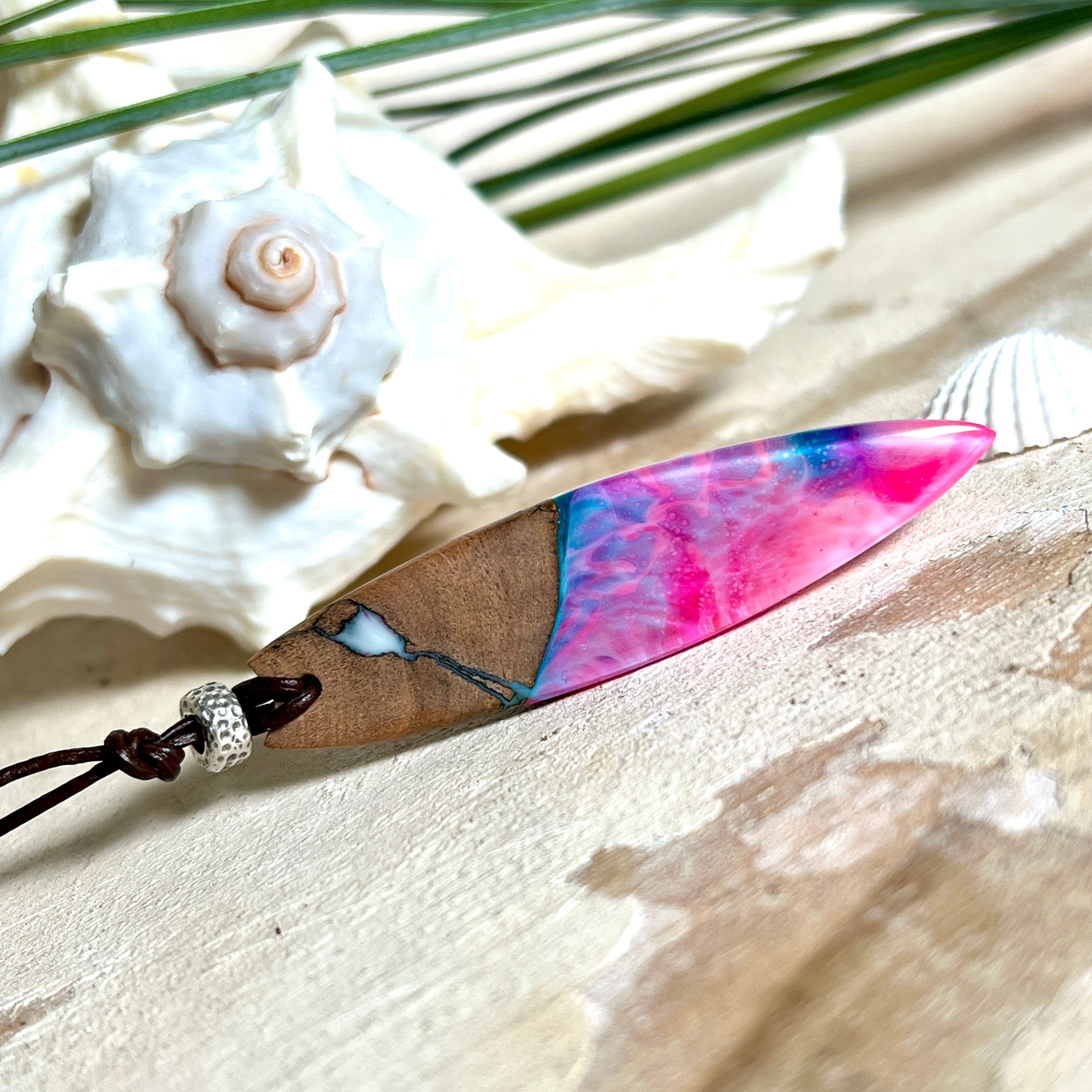 Handgefertigter Surfbrett-Anhänger aus Nussbaumholz und fluoreszierendem Epoxidharz in Pink, Blau und Weiß. Der Anhänger leuchtet zart bei Schwarzlicht und wird an einem verstellbaren Lederband (25–44 cm) mit Holzperlen getragen. Ein maritimes Unikat – ideal für Surfer und Naturliebhaber.