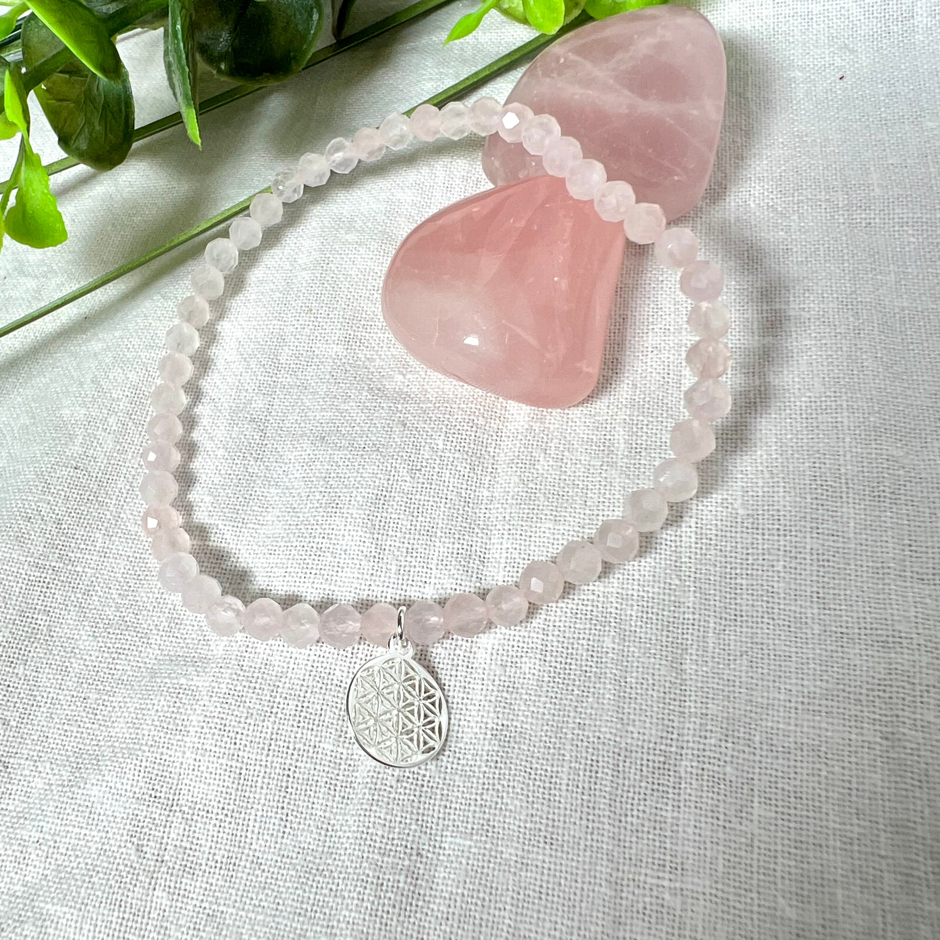 Handgefertigtes Rosenquarz-Armband mit facettierten 4mm Perlen, ergänzt durch einen filigranen Blume-des-Lebens-Anhänger aus 925er Silber. Präsentiert auf hellem Hintergrund mit Rosenquarzsteinen und goldenen Dekoelementen. Spiritueller Schmuck mit harmonisierender Wirkung.